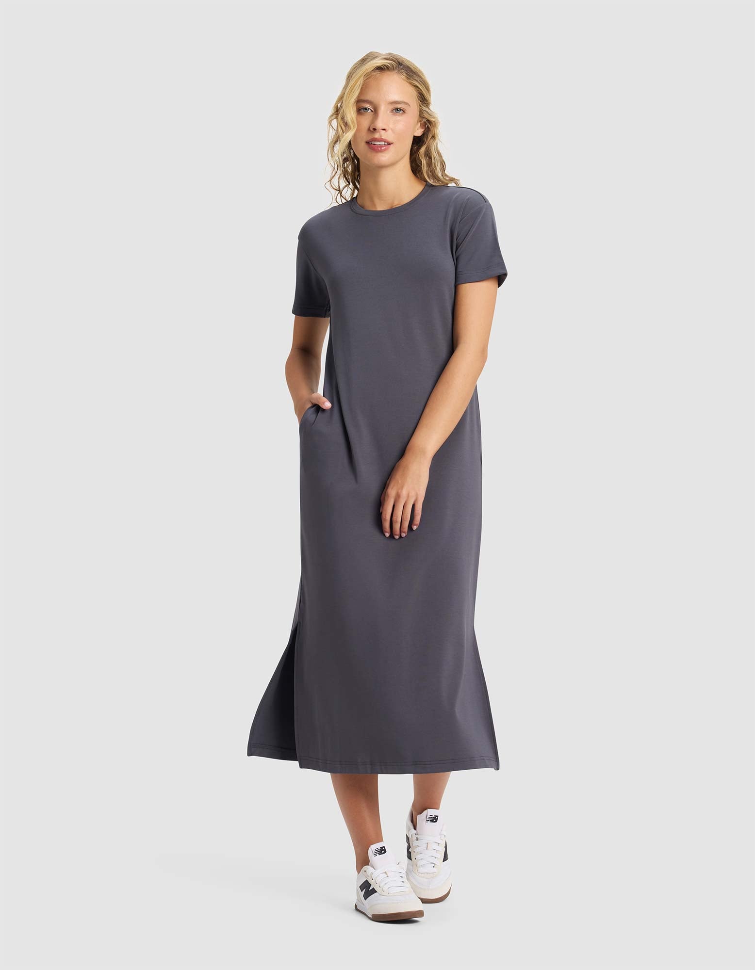 A woman smiles against a plain background, wearing the Cozy Earth Women's Brushed Bamboo Midi Dress in gray with short sleeves, side pockets, and white sneakers. She has one hand in her pocket.