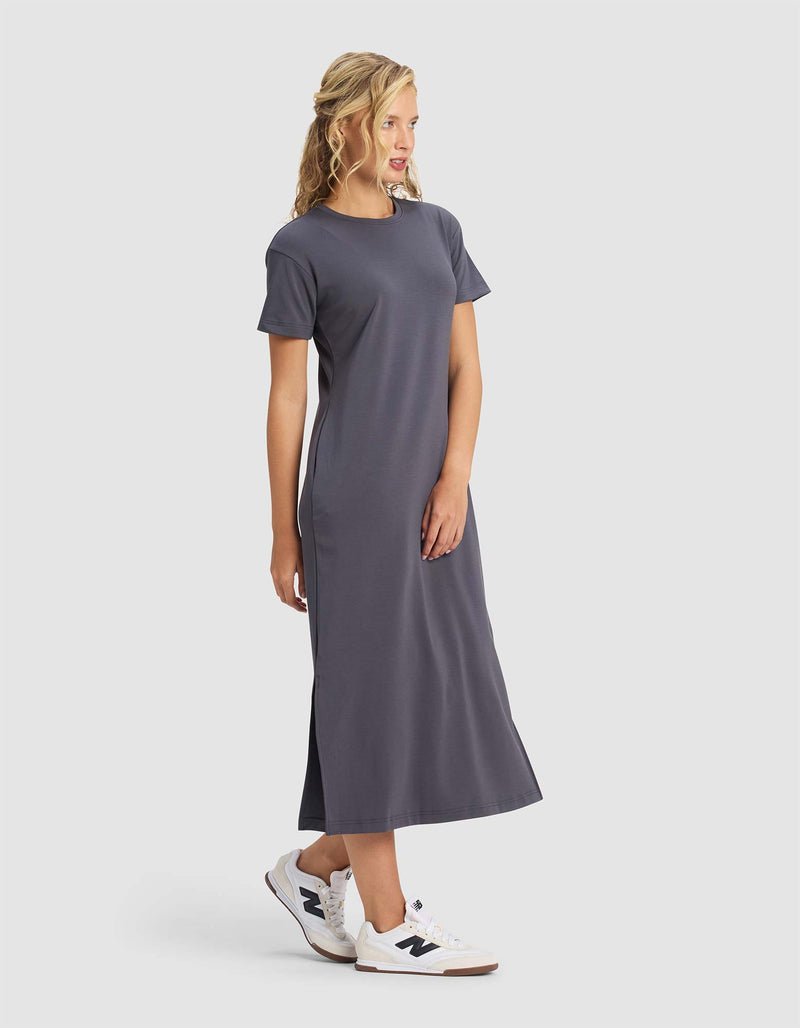 A blonde woman stands and smiles, slightly turned to the side, wearing Cozy Earth's Women's Brushed Bamboo Midi Dress in dark gray with short sleeves and white sneakers against a plain light background.