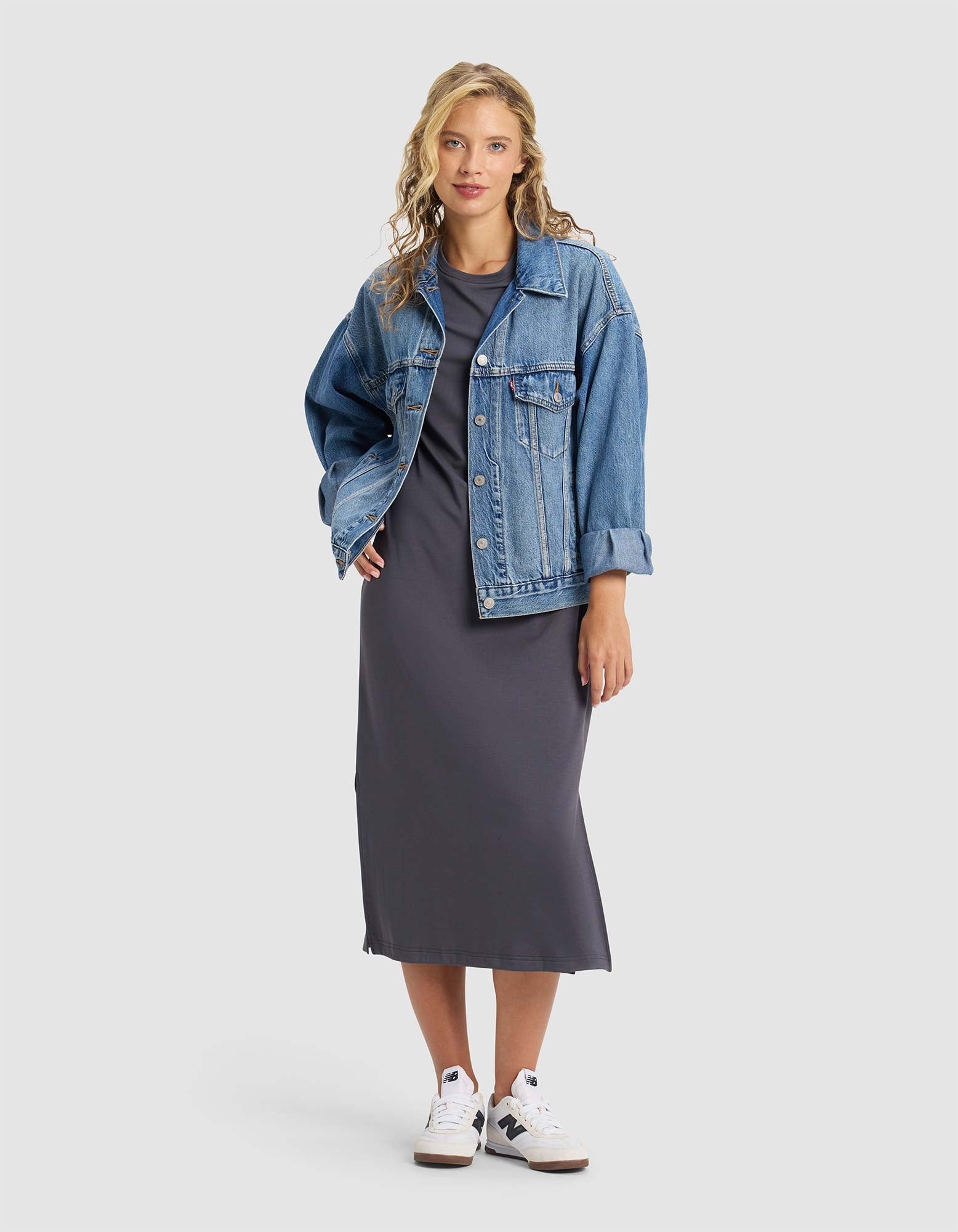 A woman with wavy blonde hair and a confident expression wears the Cozy Earth Women's Brushed Bamboo Midi Dress in dark gray, styled with a blue denim jacket and white sneakers against a plain background.|Color:Graphite