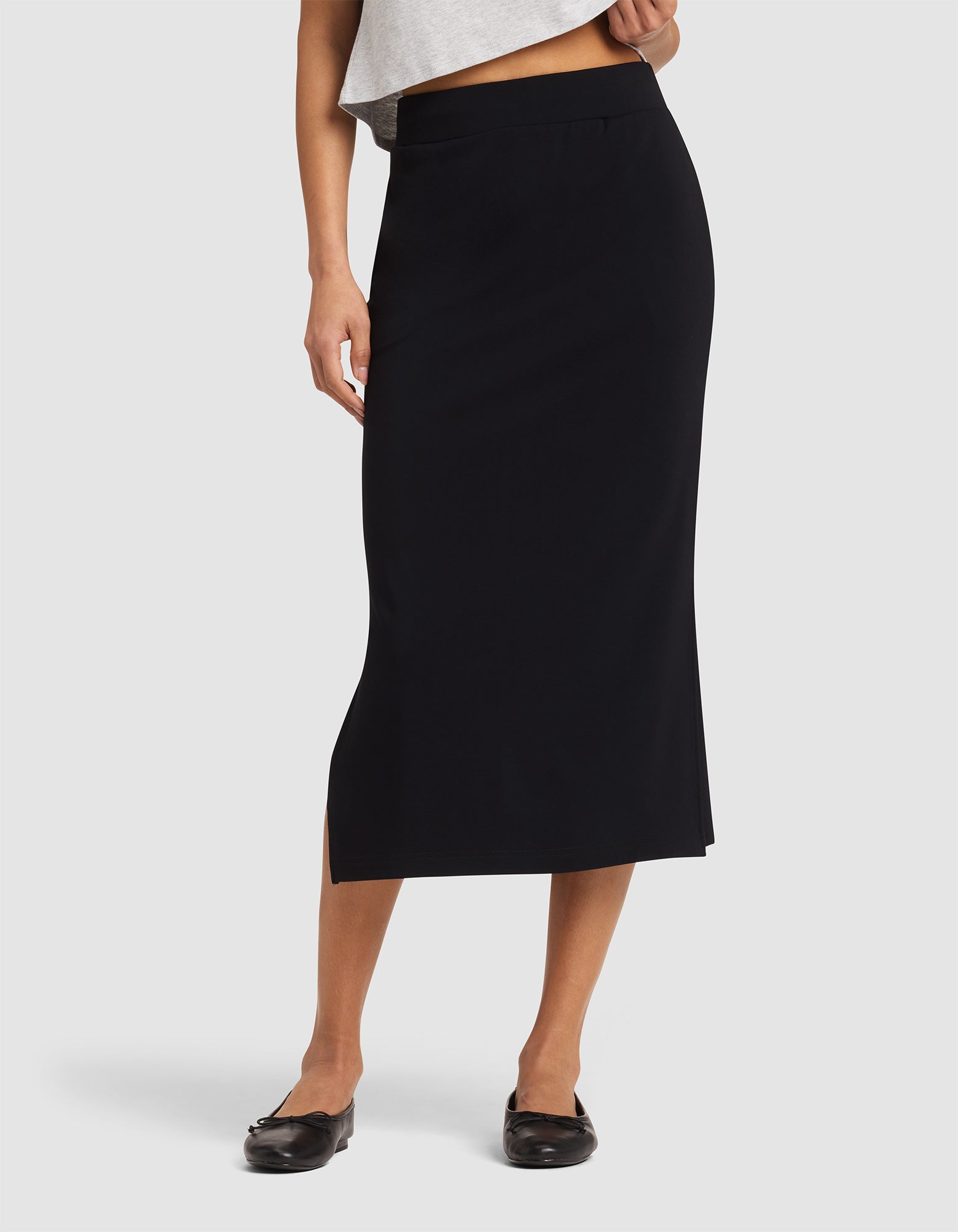 A woman wears the Cozy Earth Women's Brushed Bamboo Midi Skirt in black, high-waisted and ankle-length with a subtle side slit, paired with a gray top and black flats against a plain white background. |Color:Black