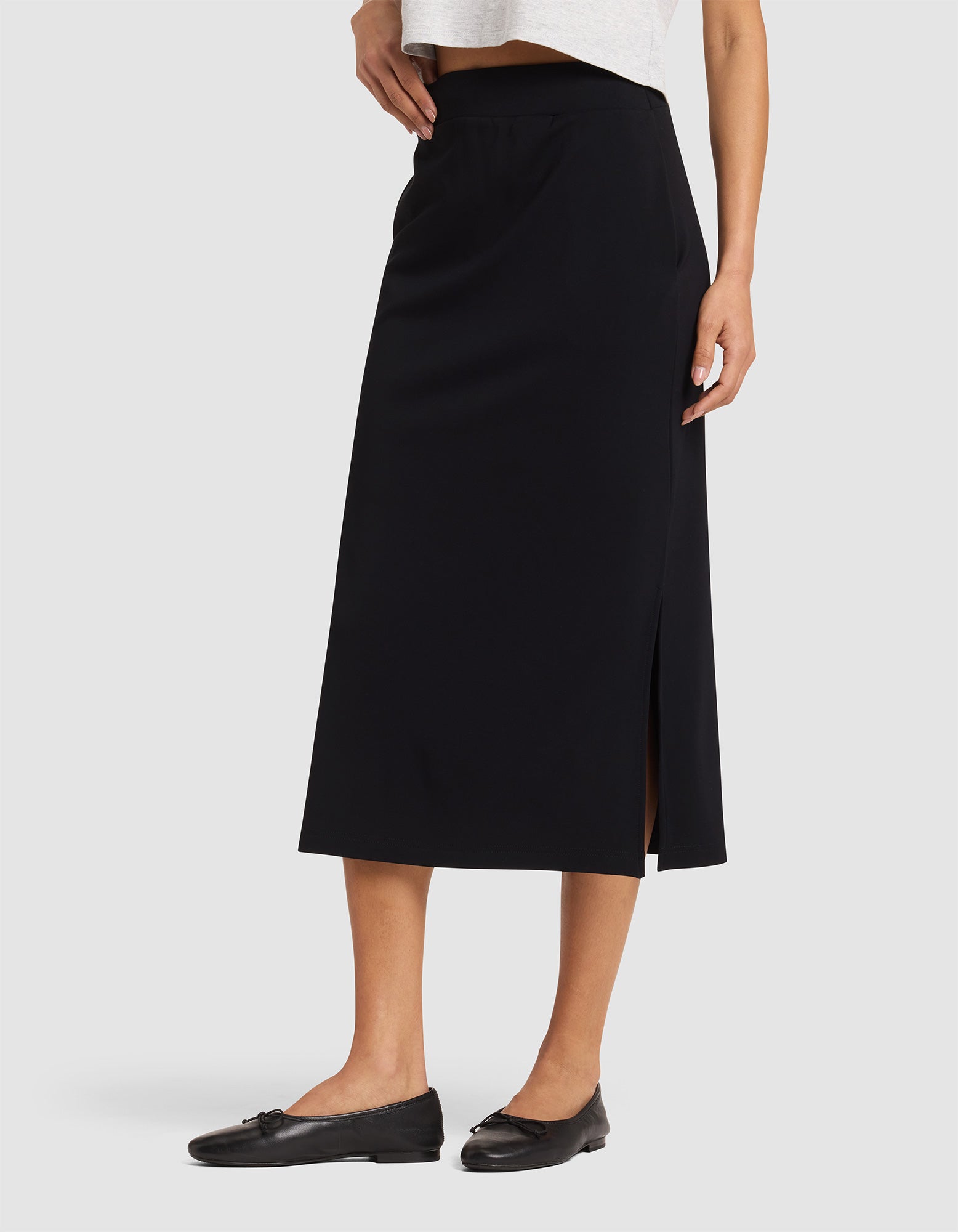 A woman wears the Cozy Earth Women's Brushed Bamboo Midi Skirt—black, knee-length with a side slit—paired with a white top and black flats against a plain light background. |Color:Black