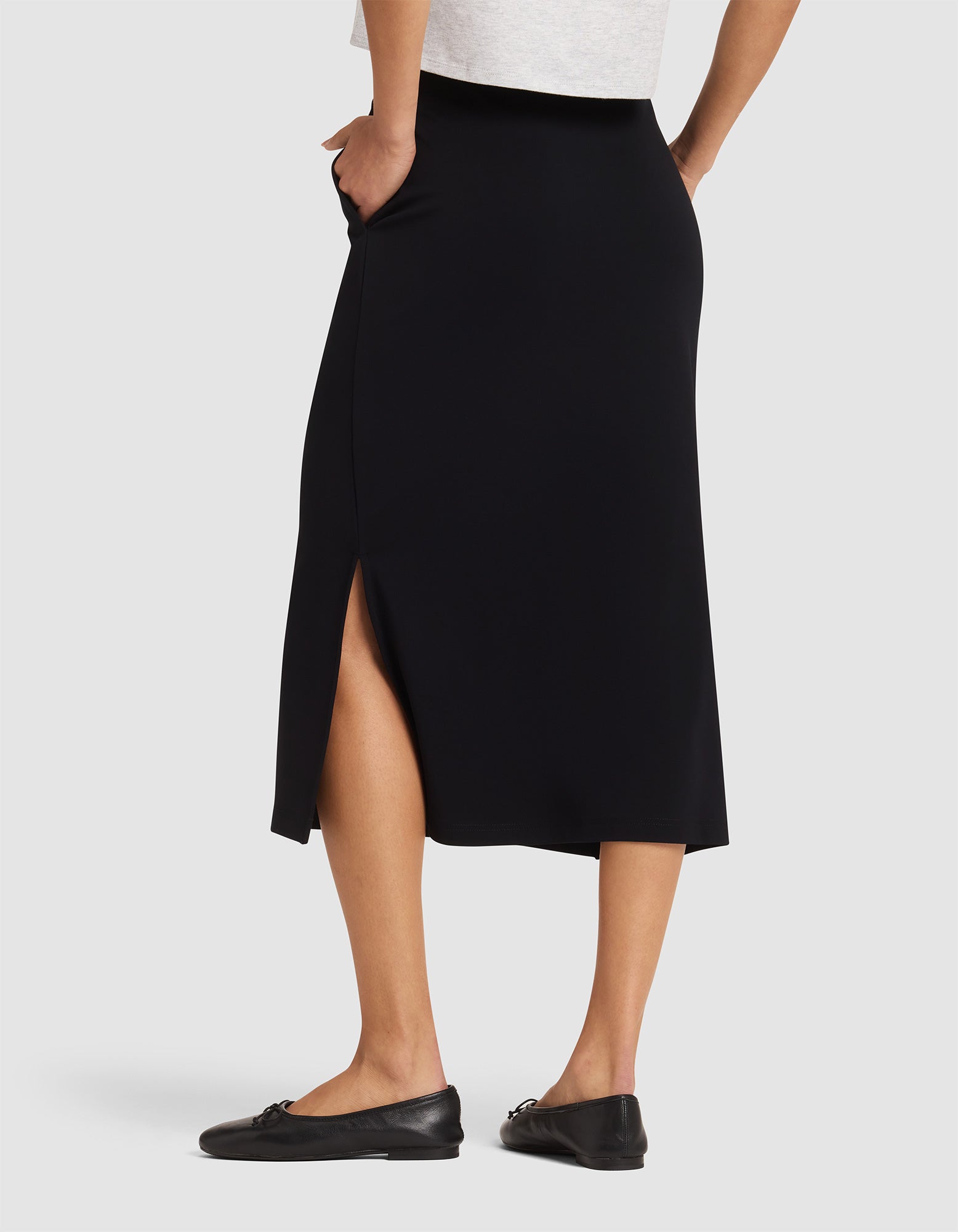Model wears Cozy Earth's Women's Brushed Bamboo Midi Skirt, featuring a side slit, paired with black flats and a light top. She stands hands in pockets, showcasing the skirt’s back and side views. 