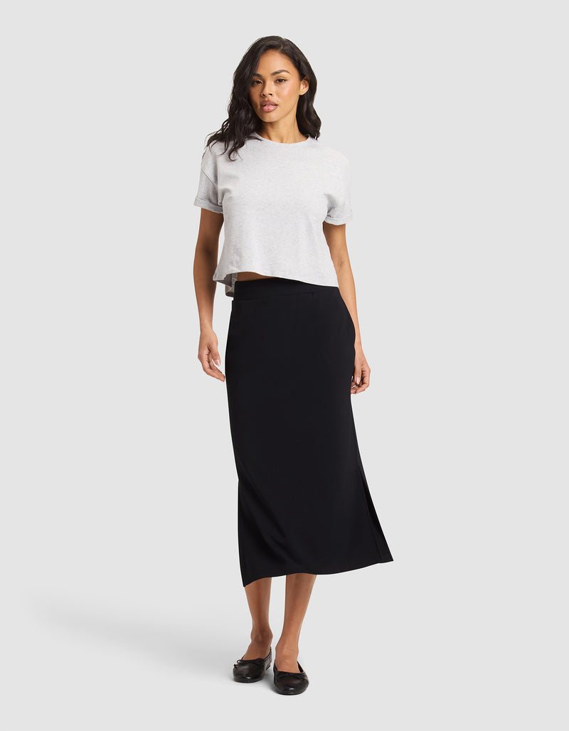 A woman with wavy dark hair and a confident stance wears the Cozy Earth Women's Brushed Bamboo Midi Skirt in black, paired with a light gray short-sleeve top and black flats, standing against a plain background.