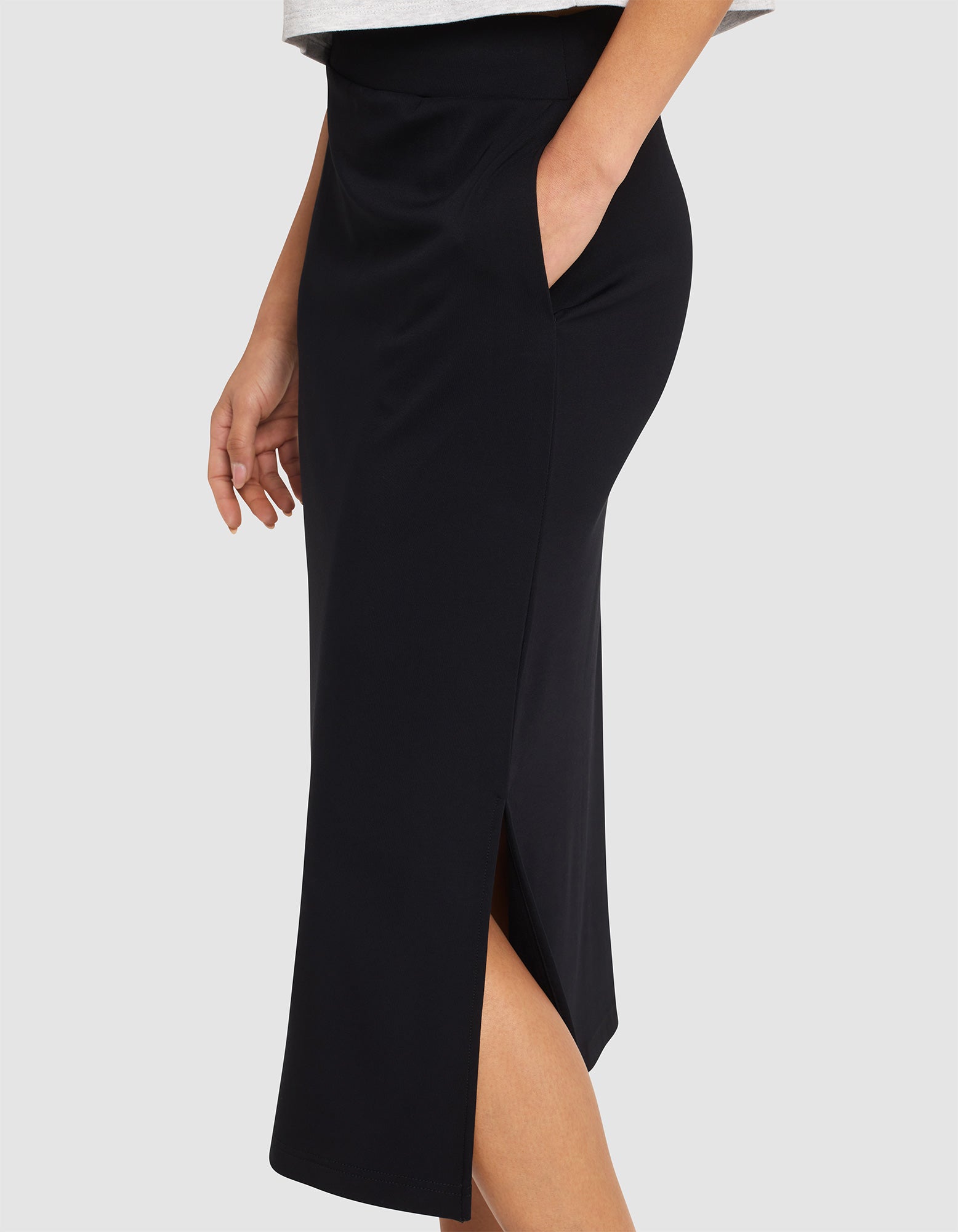 A woman models the Cozy Earth Women's Brushed Bamboo Midi Skirt—black, knee-length with a side slit and pockets—standing with one hand in her pocket against a plain light background.