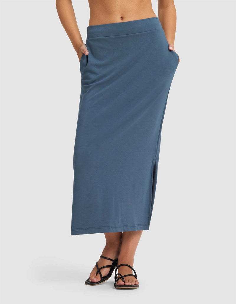 A person wears the Cozy Earth Women's Brushed Bamboo Midi Skirt—a high-waisted, ankle-length blue skirt with side pockets and a side slit—paired with black strappy sandals against a plain white background.