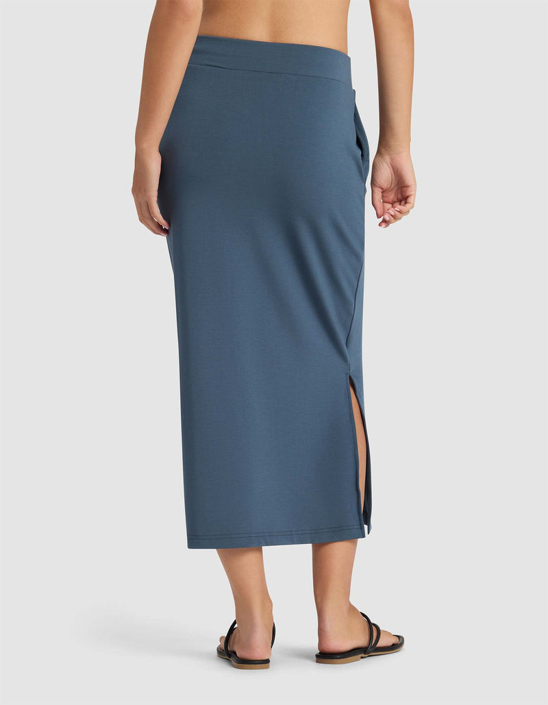 A woman is shown from the back wearing Cozy Earth’s Women’s Brushed Bamboo Midi Skirt in blue with a side slit, paired with black sandals, set against a plain white background.
