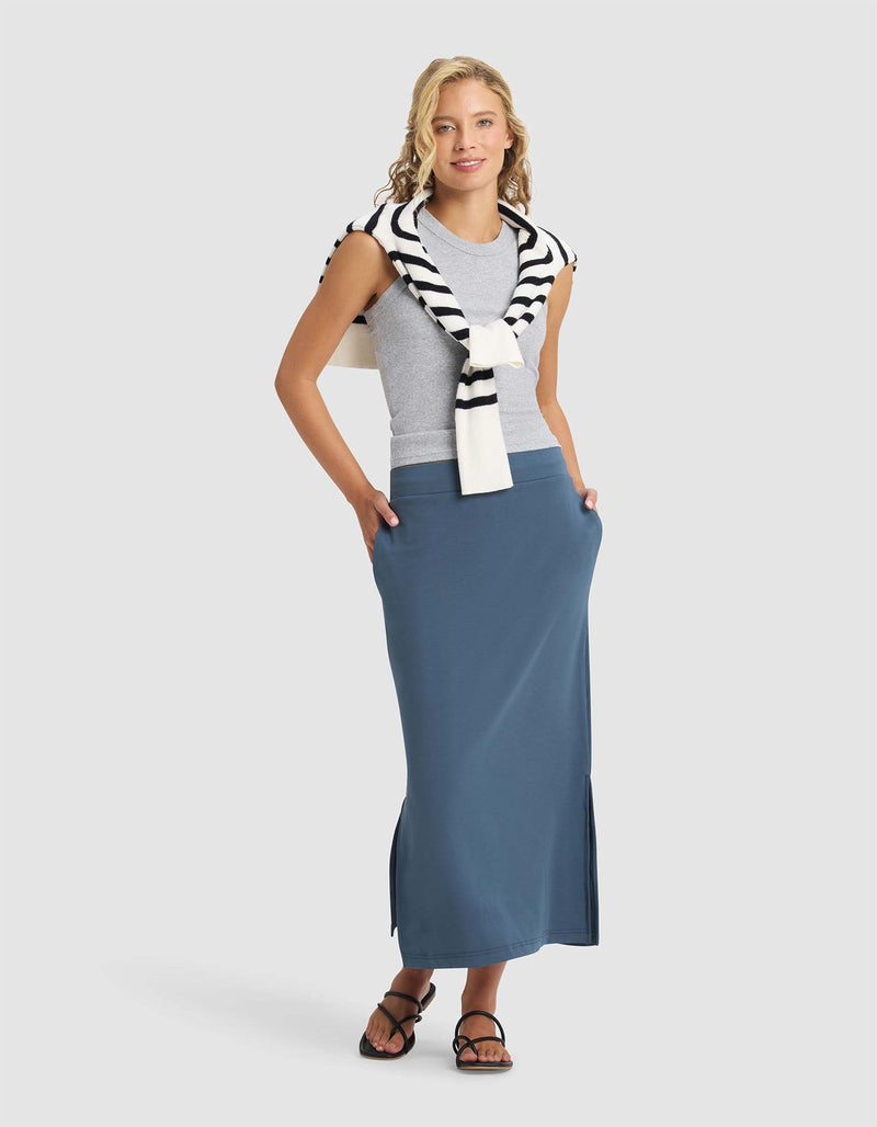 A woman smiles with hands in her pockets, wearing a grey sleeveless top, black sandals, and Cozy Earth's Women's Brushed Bamboo Midi Skirt in blue. A white sweater with black stripes is draped over her shoulders.
