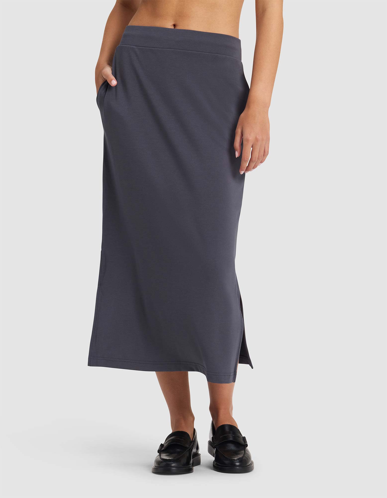 A person is wearing the Cozy Earth Women's Brushed Bamboo Midi Skirt in dark gray, featuring a high waist, side pockets, and small side slits, paired with black loafers. The upper body and face are not visible.