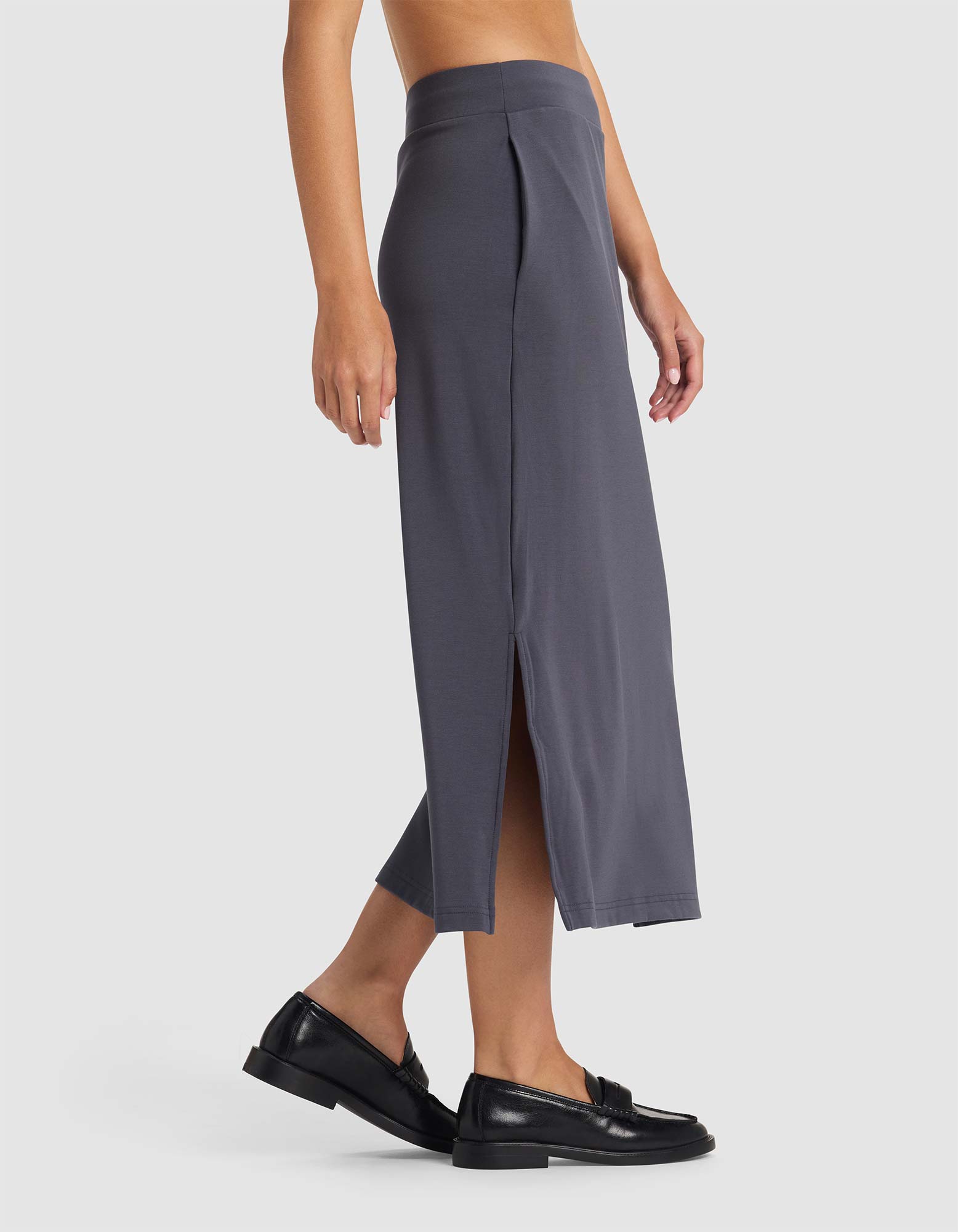 A person is seen from the side wearing Cozy Earth’s Women’s Brushed Bamboo Midi Skirt in gray, featuring a high waist and side slit, paired with black loafers against a light gray background.