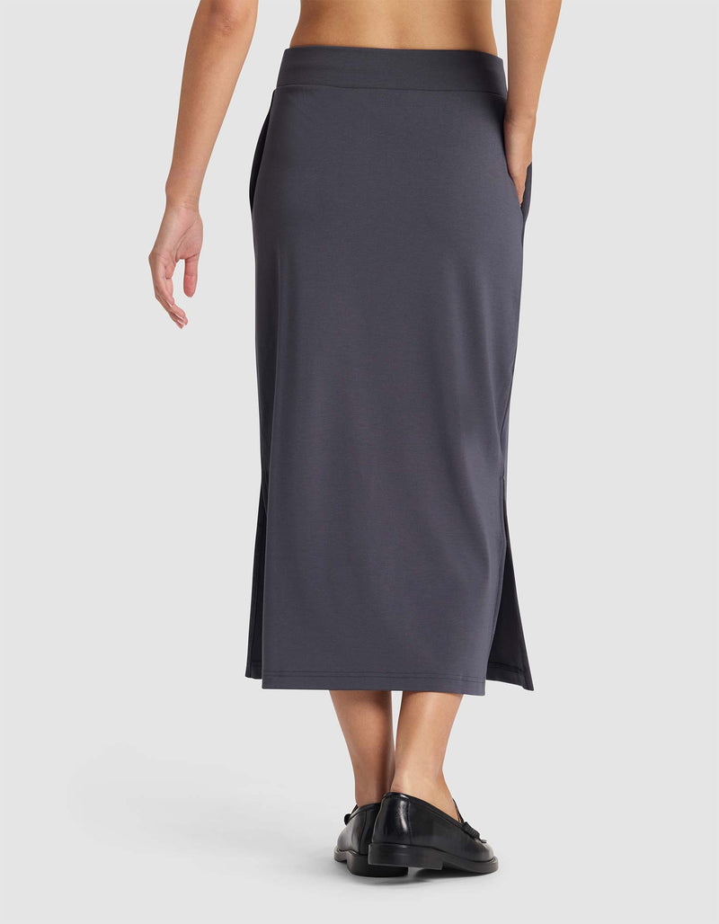 A person is shown from the back wearing the Cozy Earth Women's Brushed Bamboo Midi Skirt in dark gray with side slits and black shoes, set against a plain white background.