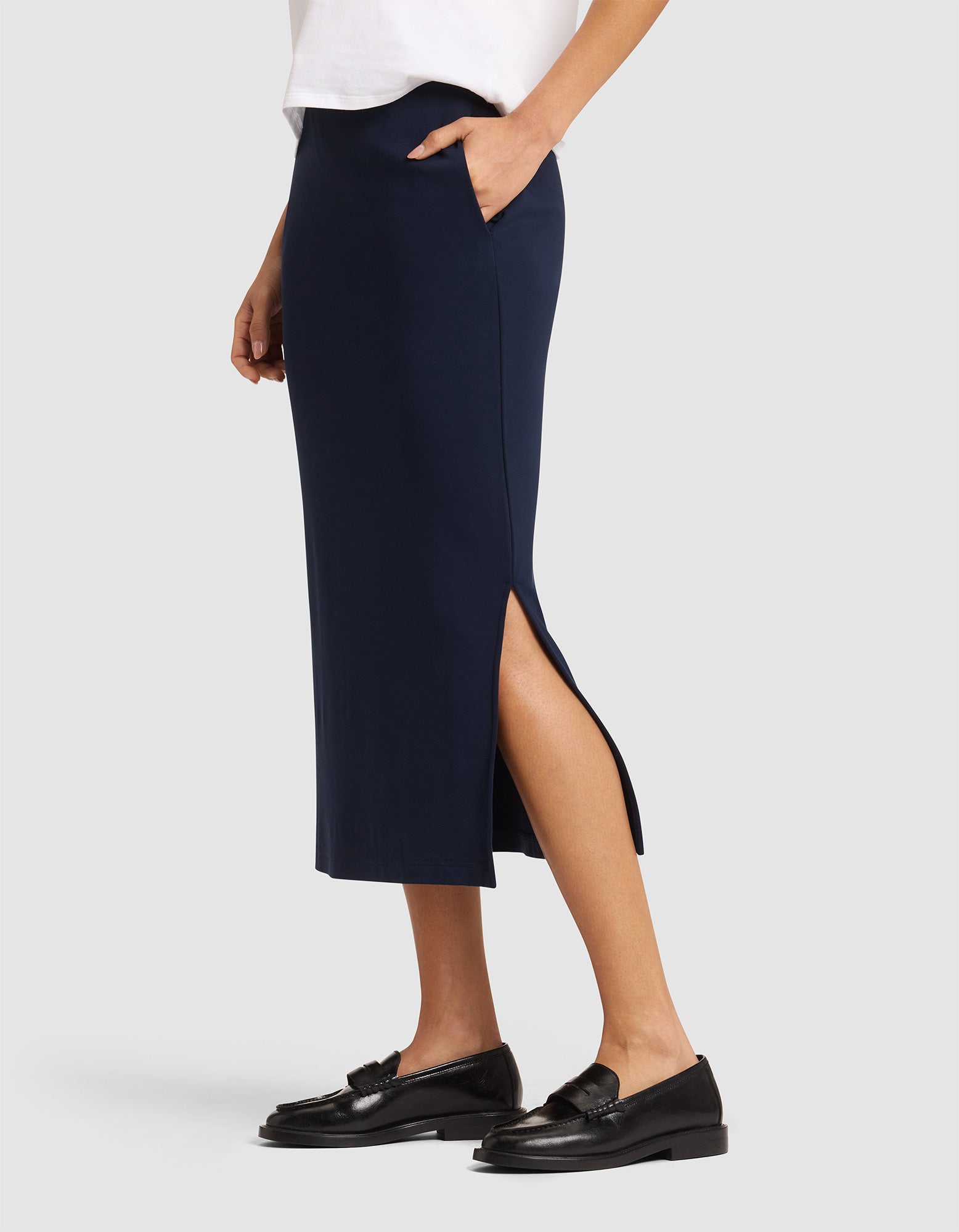 A person wearing a white top and Cozy Earth's Women's Brushed Bamboo Midi Skirt in navy blue with a side slit and pockets, paired with black loafers, stands against a plain light background.  
