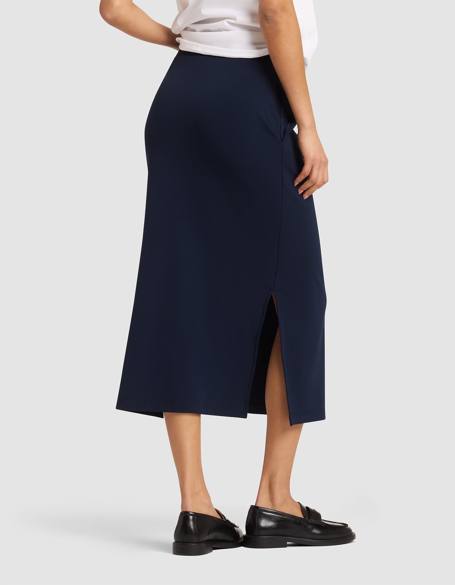A woman is shown from the back wearing the Cozy Earth Women's Brushed Bamboo Midi Skirt in navy blue with a side slit, paired with a white top and black loafers, against a plain light gray background.  |Color:Navy