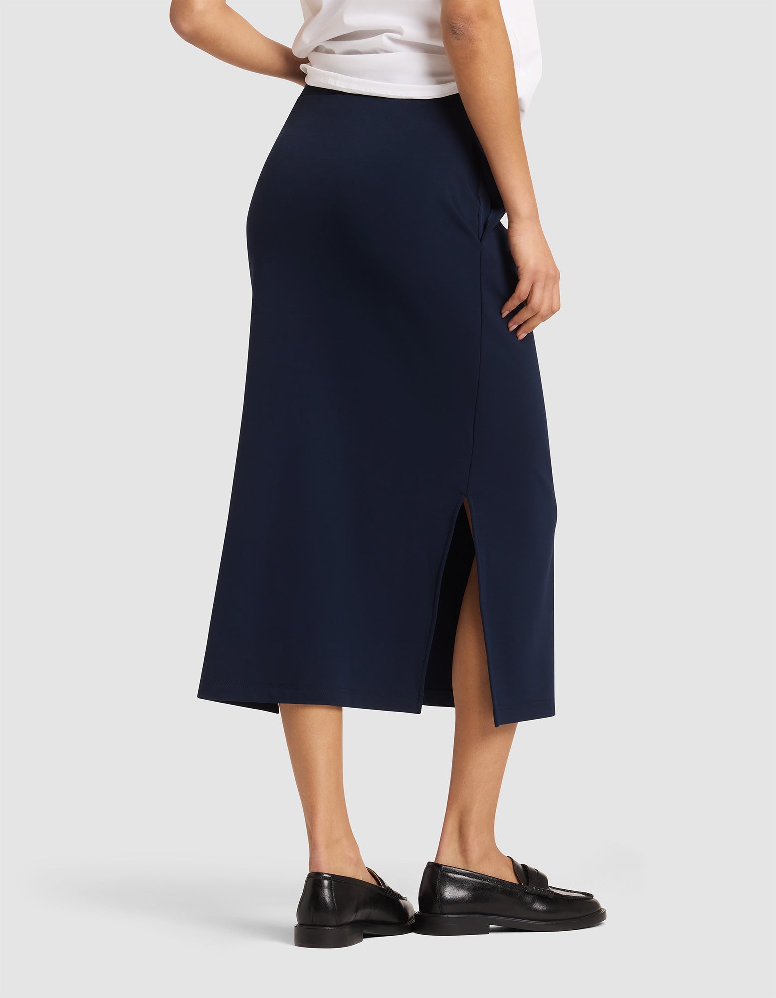 A woman is shown from the back wearing the Cozy Earth Women's Brushed Bamboo Midi Skirt in navy blue with a side slit, paired with a white top and black loafers, against a plain light gray background.  