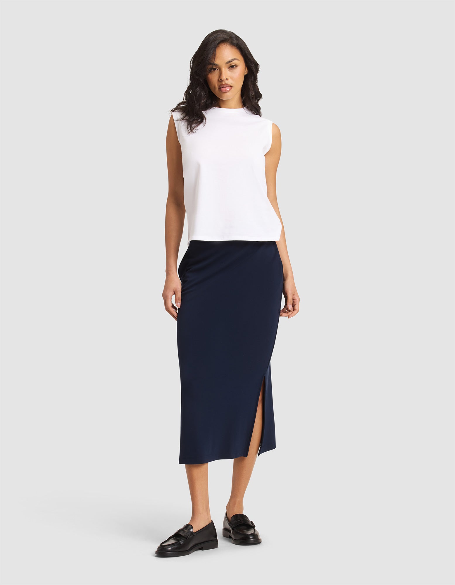 A woman stands against a plain background wearing a sleeveless white top, black loafers, and the Cozy Earth Women's Brushed Bamboo Midi Skirt in navy blue with a side slit.  