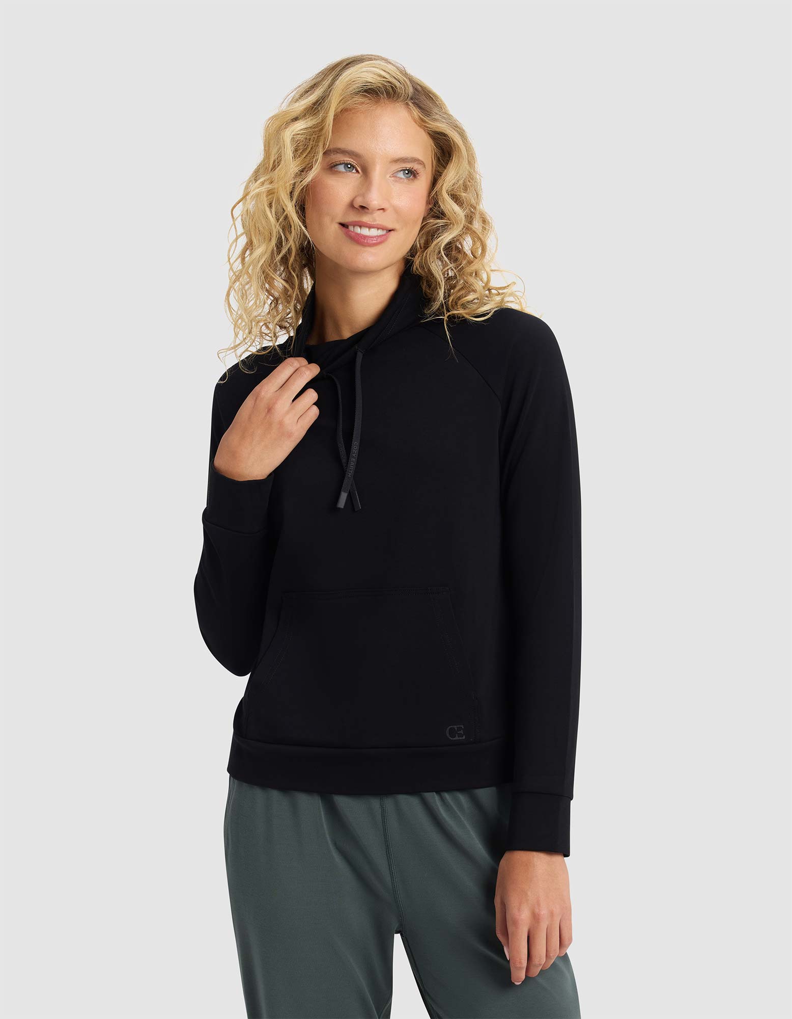A smiling woman with blonde curly hair is wearing the Cozy Earth Women's Brushed Bamboo Mock Neck, paired with green pants, standing against a light gray background and looking to the side. |Color:Black
