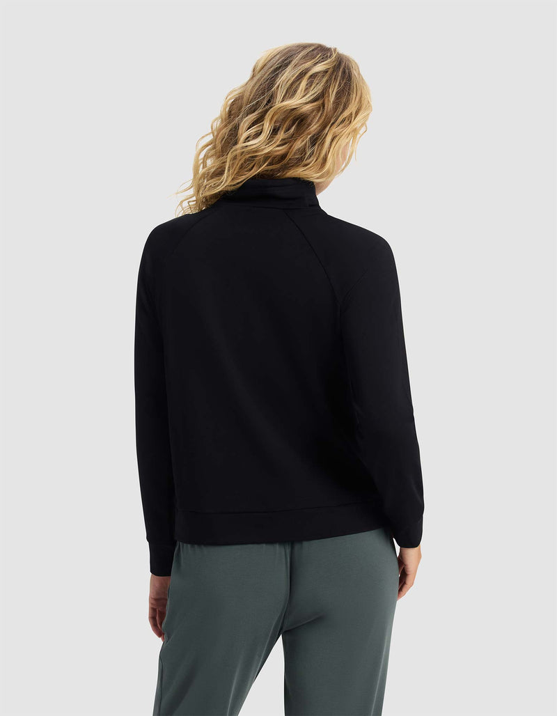 A woman with wavy blonde hair is shown from the back, wearing the Cozy Earth Women's Brushed Bamboo Mock Neck in black, paired with green pants against a plain light gray background.