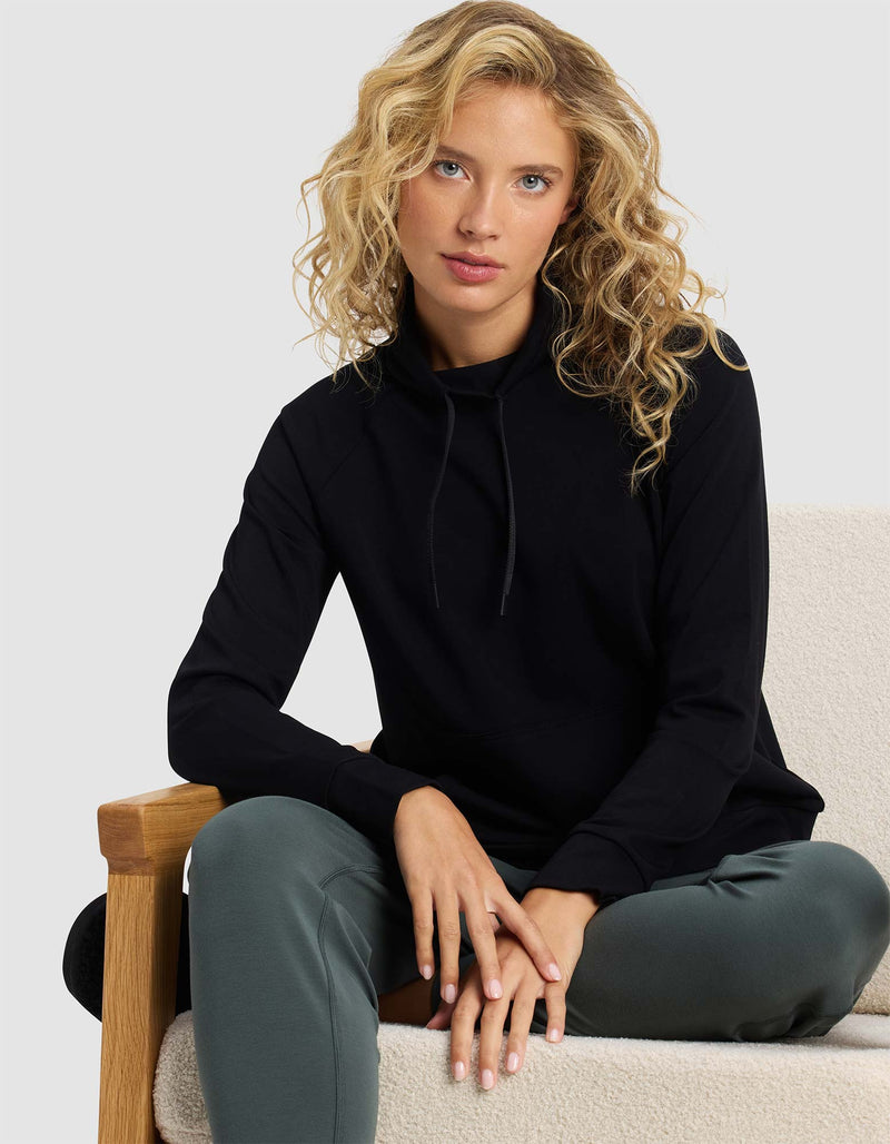 A woman with wavy blond hair sits on a light upholstered chair, facing forward with her hands on her knees, wearing a black Cozy Earth Women's Brushed Bamboo Mock Neck top and dark green pants.