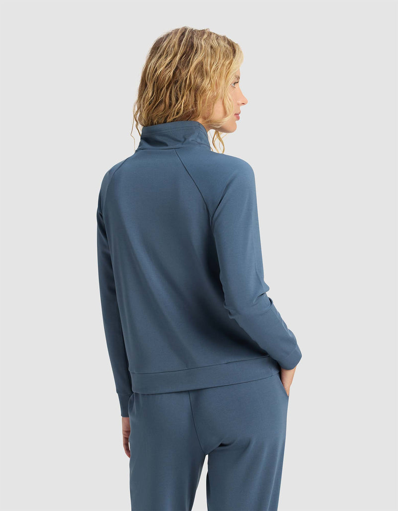 A woman with wavy blonde hair faces away, wearing the Cozy Earth Women's Brushed Bamboo Mock Neck and matching blue pants. Her hands are in her pockets against a plain light gray background.