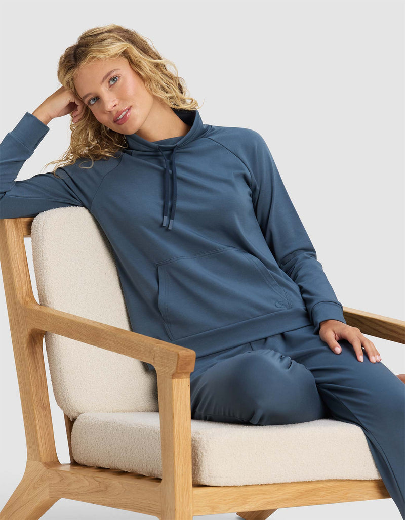 A woman with curly blonde hair, wearing the Cozy Earth Women's Brushed Bamboo Mock Neck in blue, sits relaxed on a wooden armchair with cream cushions, resting her head on one hand and smiling softly.