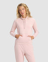 A woman with wavy blonde hair wears the Cozy Earth Women's Brushed Bamboo Mock Neck in light pink, paired with matching pants, standing with her hands in the hoodie pocket against a plain light gray background.|Color:Dusty Rose