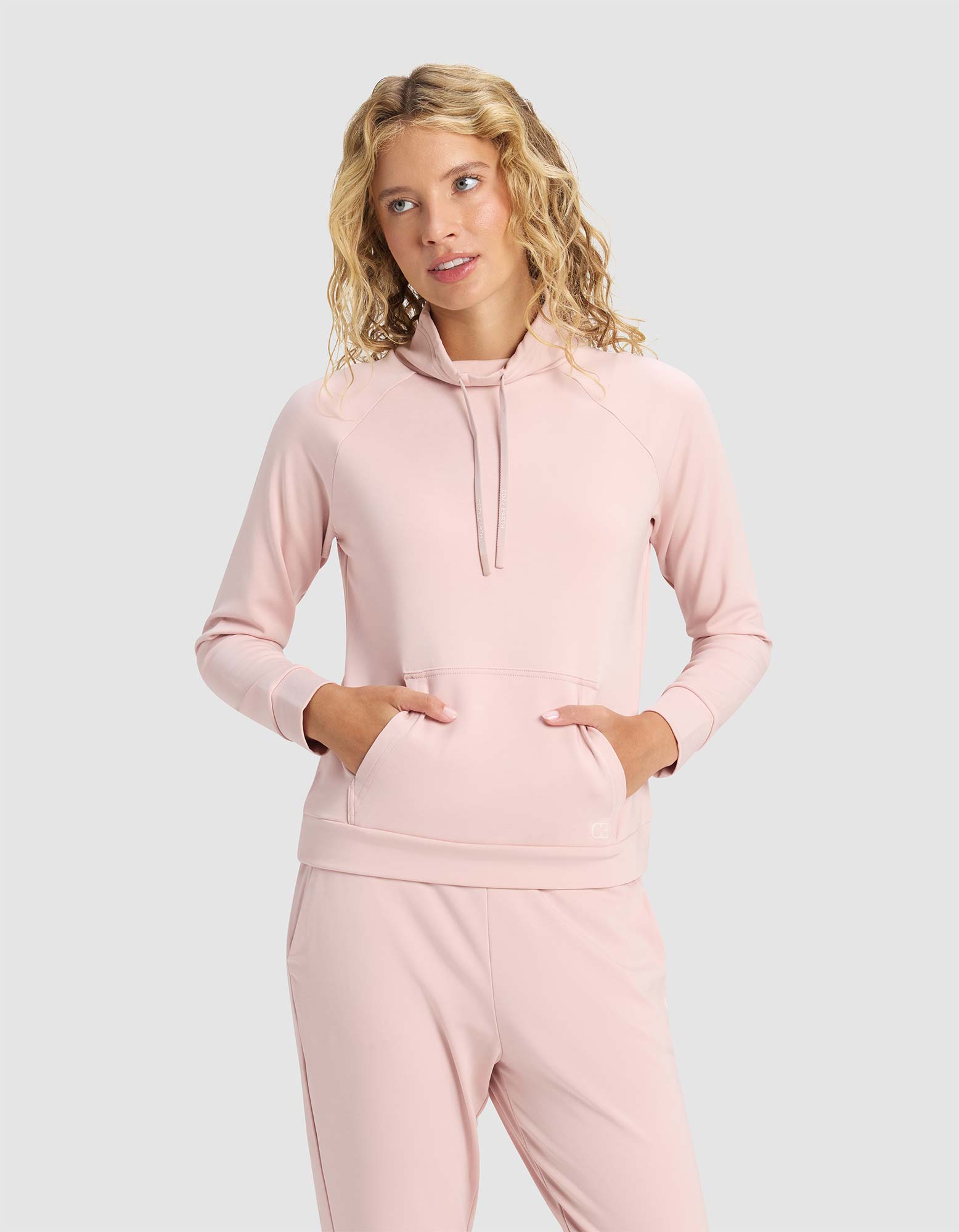 A woman with wavy blonde hair wears the Cozy Earth Women's Brushed Bamboo Mock Neck in light pink, paired with matching pants, standing with her hands in the hoodie pocket against a plain light gray background.