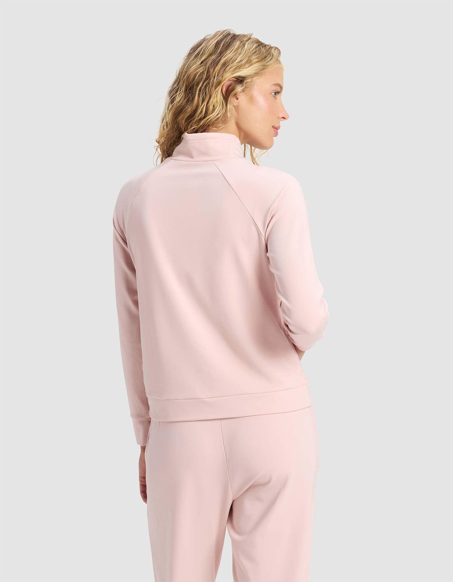 A woman with wavy blonde hair wears the Cozy Earth Women's Brushed Bamboo Mock Neck in light pink, standing against a plain light gray background with her head turned slightly to the side.