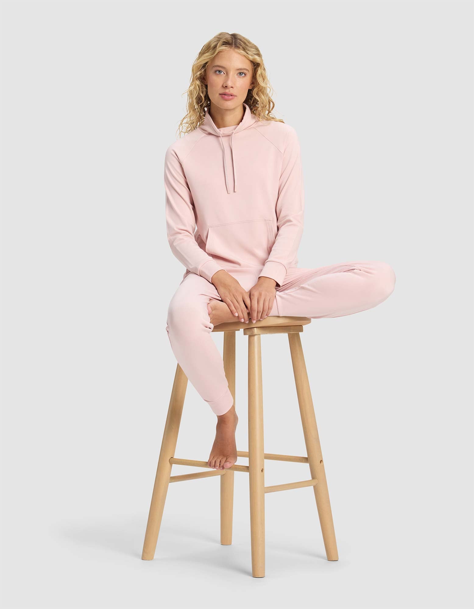 A woman wearing the Cozy Earth Women's Brushed Bamboo Mock Neck sits barefoot on a wooden stool, one leg bent and the other hanging down, against a plain light gray background.