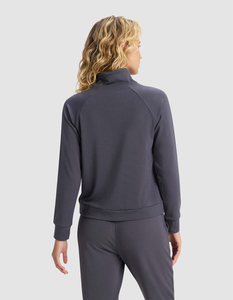 A person with wavy blonde hair models the back of Cozy Earth's Women's Brushed Bamboo Mock Neck in dark gray, along with matching pants, standing against a plain light background.