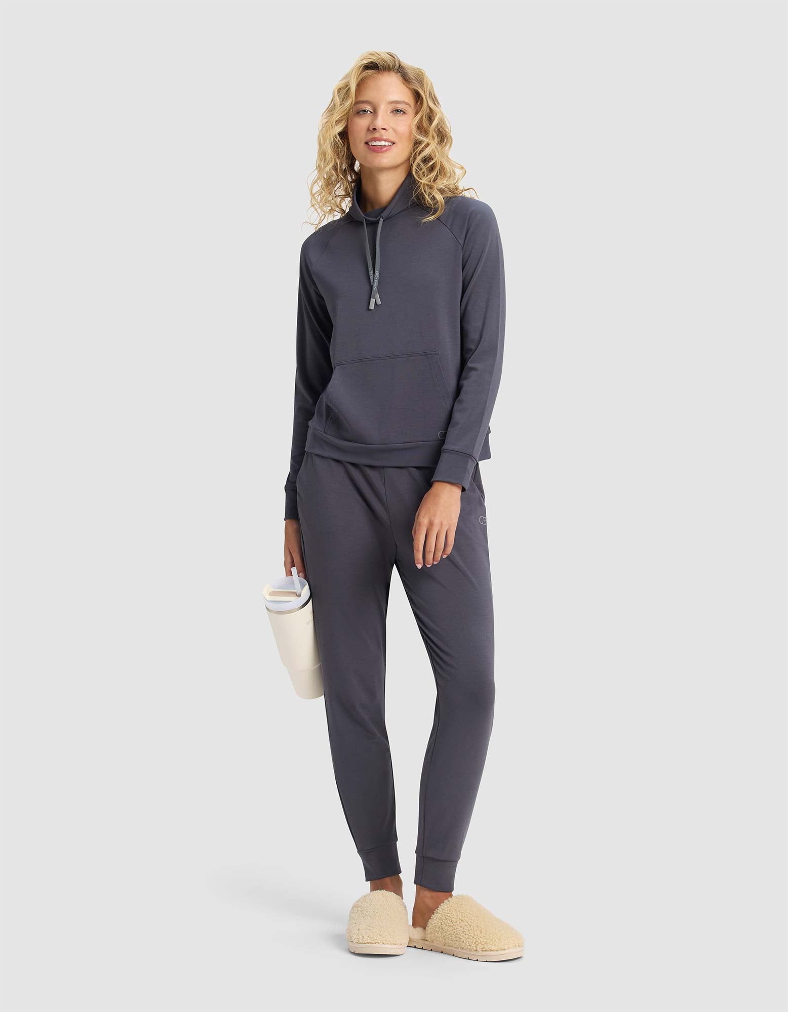 A smiling woman wears the Cozy Earth Women's Brushed Bamboo Mock Neck in dark gray with tan slippers, holding a white water bottle against a plain light background.