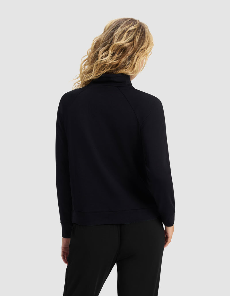 A woman with wavy blonde hair is seen from behind, wearing Cozy Earth's Women's Brushed Bamboo Mock Neck & Jogger Set in black, against a light gray background.