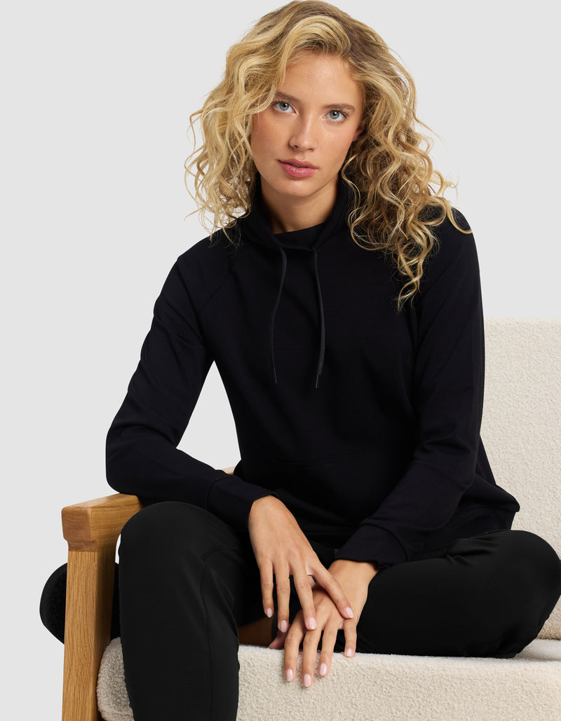 A woman with curly blonde hair wears the Cozy Earth Women's Brushed Bamboo Mock Neck & Jogger Set, sitting on a light cushioned chair with one arm resting on the wooden armrest.