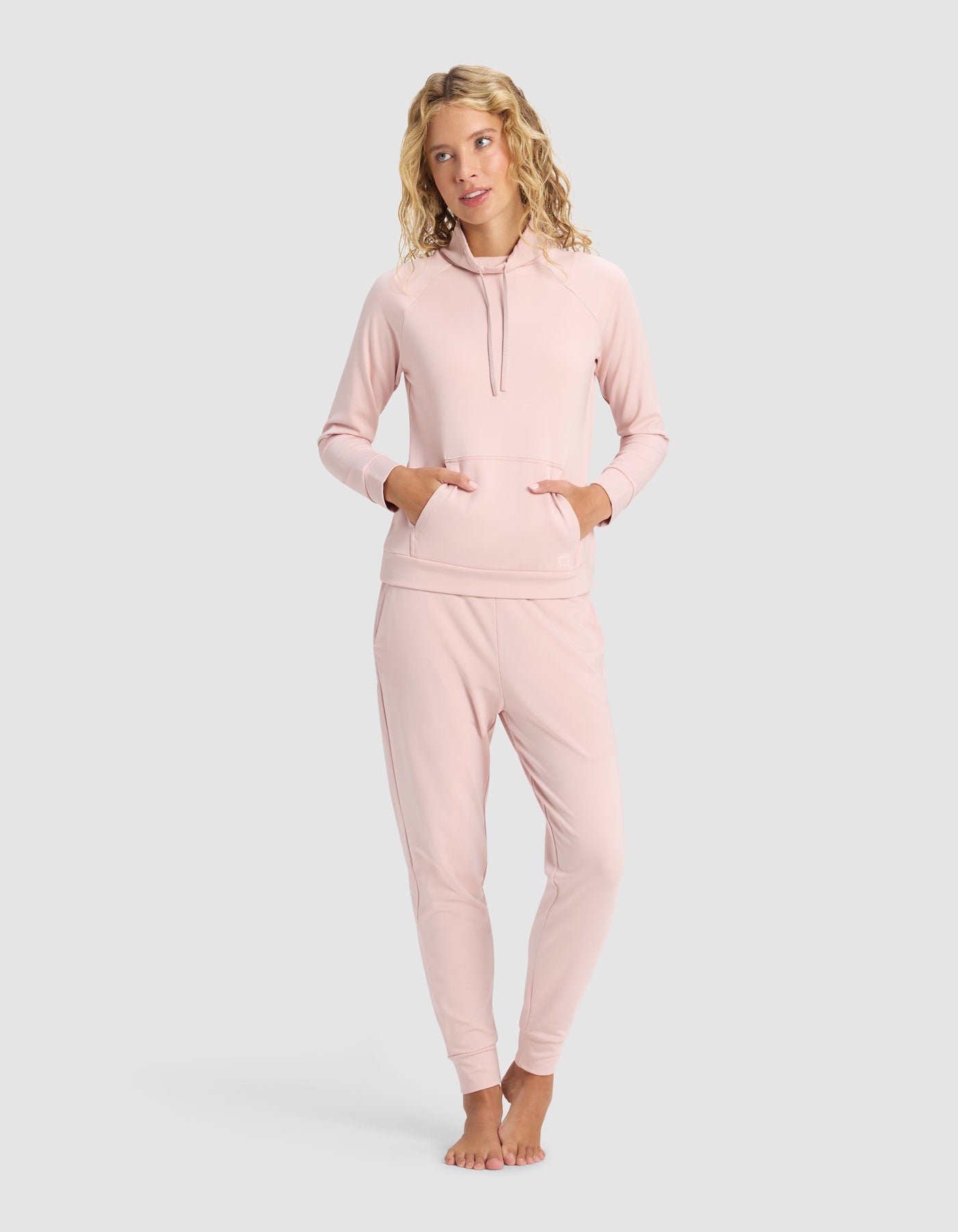 A woman with blonde wavy hair stands barefoot against a light gray background, wearing the Cozy Earth Women's Brushed Bamboo Mock Neck & Jogger Set in light pink, featuring front pockets and a relaxed fit.
