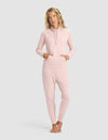 A woman with blonde wavy hair stands barefoot against a light gray background, wearing the Cozy Earth Women's Brushed Bamboo Mock Neck & Jogger Set in light pink, featuring front pockets and a relaxed fit.|Color:Dusty Rose
