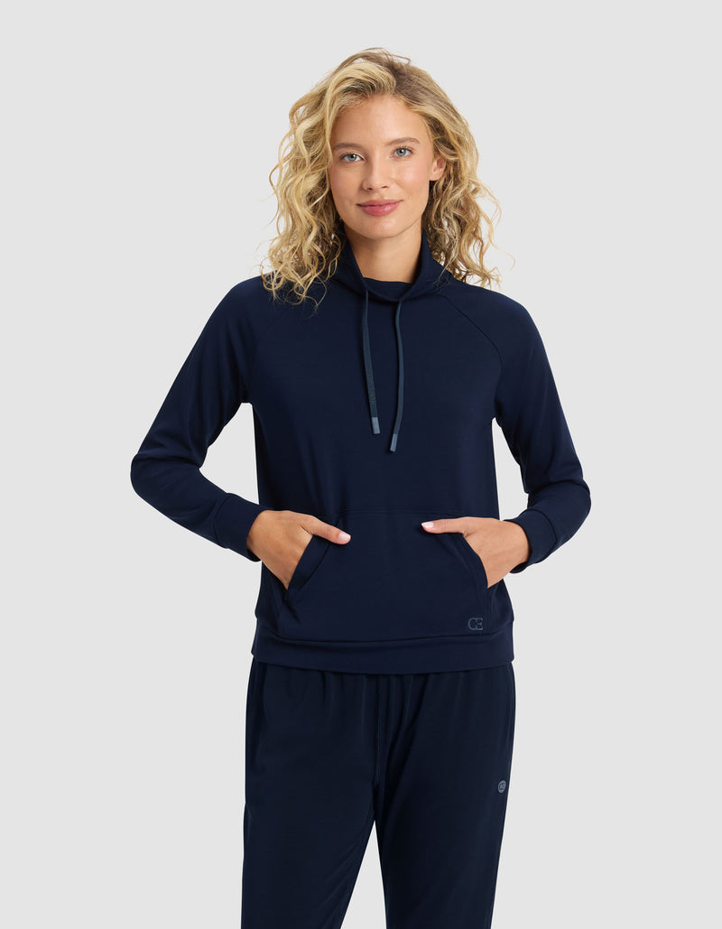 A blonde woman smiles with her hands in her pockets, wearing the Cozy Earth Women's Brushed Bamboo Mock Neck & Jogger Set in navy blue, standing against a plain light gray background.
