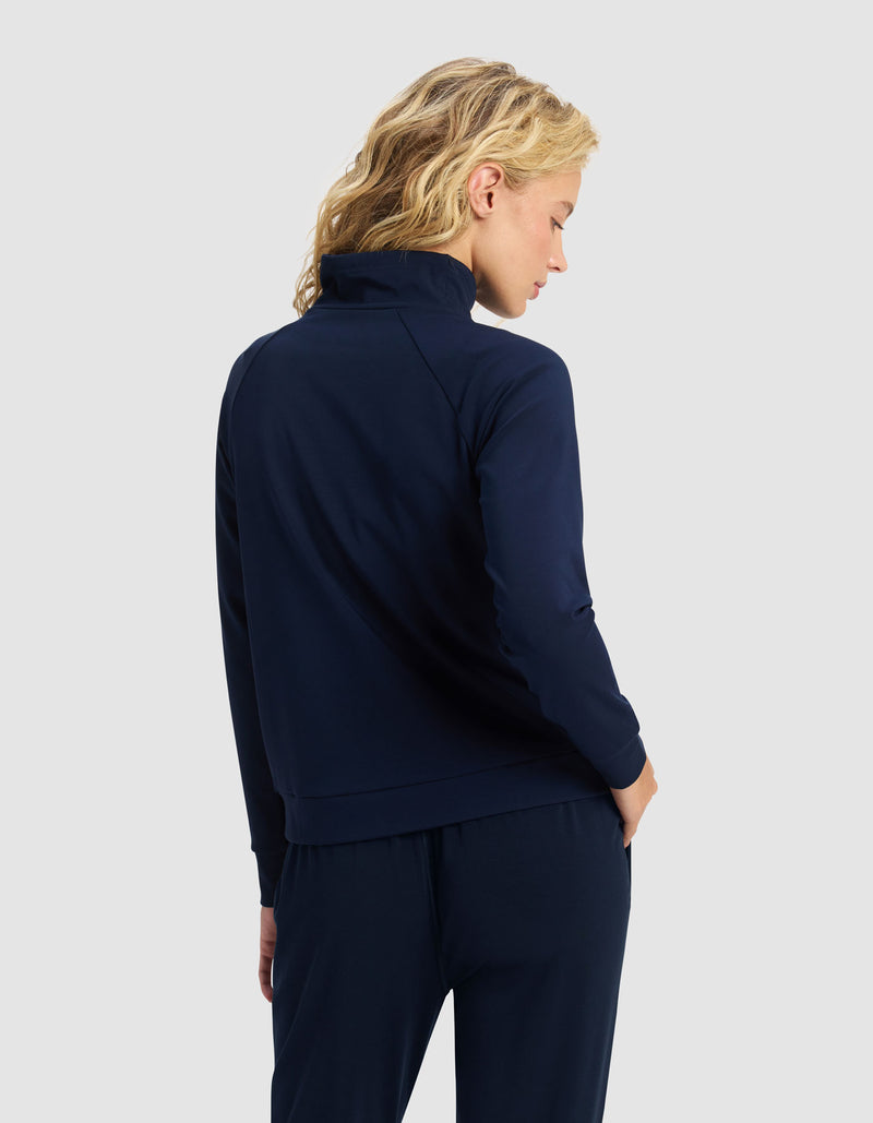 A woman with blonde, wavy hair is seen from behind wearing the Cozy Earth Women's Brushed Bamboo Mock Neck & Jogger Set in navy blue, photographed against a plain light gray background.