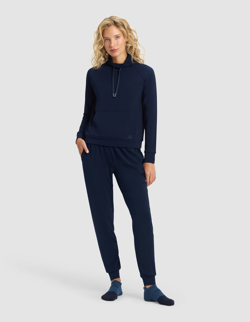 A woman with blonde, curly hair stands against a light gray background, wearing the Cozy Earth Women's Brushed Bamboo Mock Neck & Jogger Set in navy blue, paired with matching socks. She has one hand in her pocket and looks relaxed.
