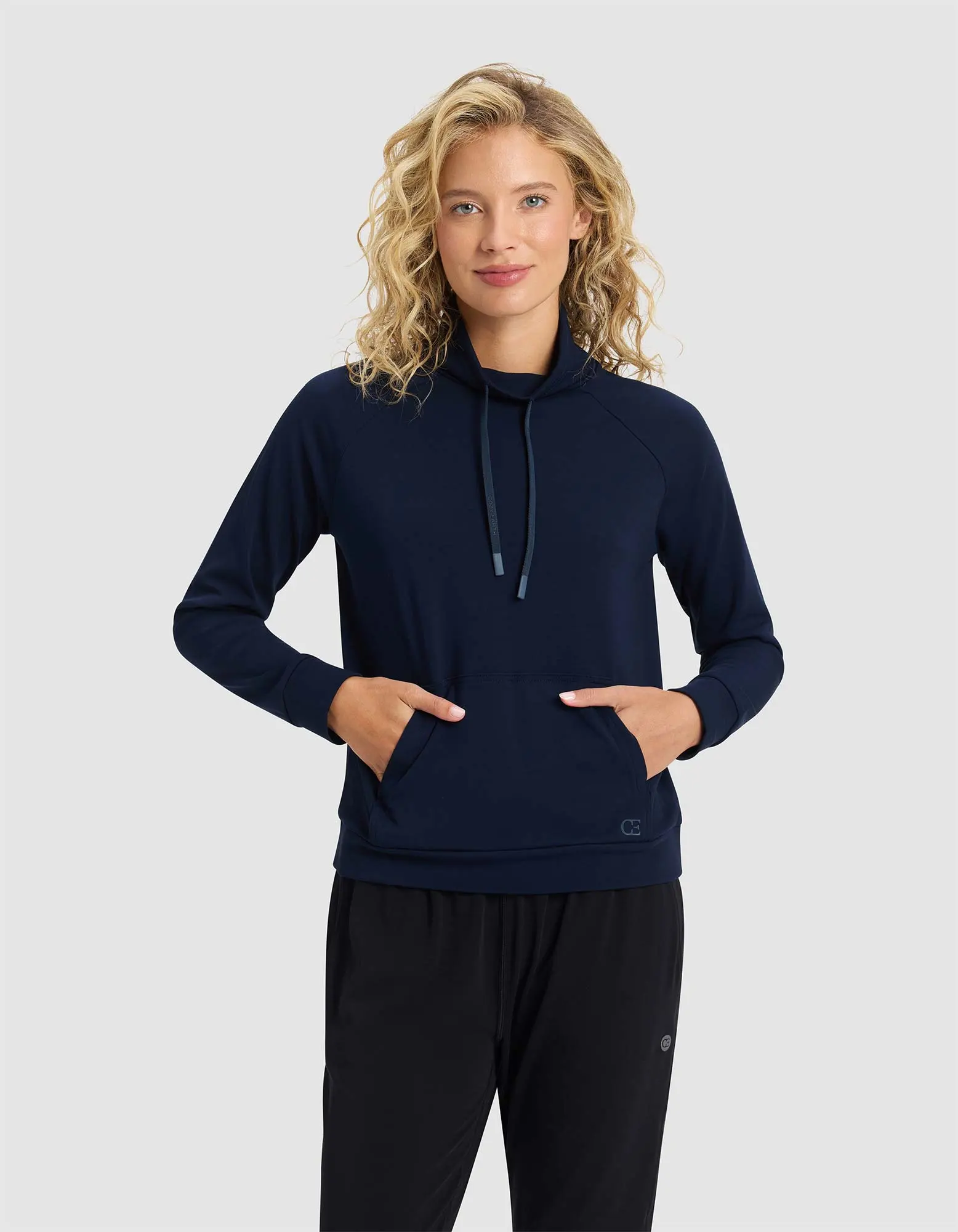 A woman with curly blonde hair models the Cozy Earth Women's Brushed Bamboo Mock Neck, a navy long-sleeve pullover with a high collar and drawstrings, standing with hands in front pockets against a plain light gray background.|Color:Navy