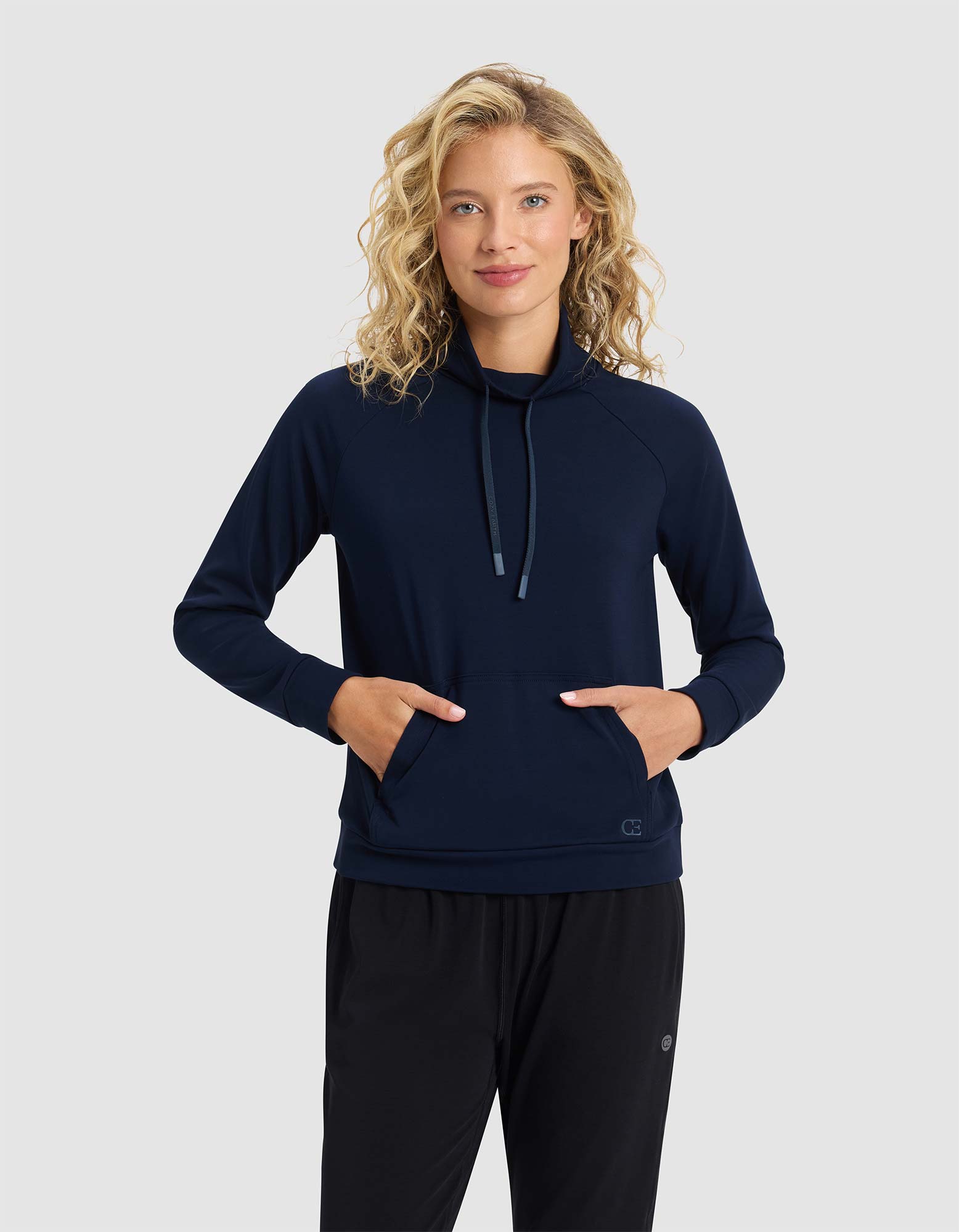 A woman with curly blonde hair models the Cozy Earth Women's Brushed Bamboo Mock Neck, a navy long-sleeve pullover with a high collar and drawstrings, standing with hands in front pockets against a plain light gray background.|Color:Navy