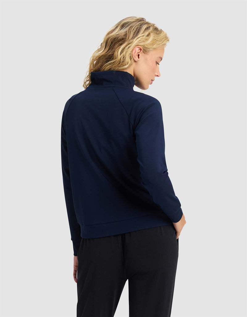 A woman with blonde hair stands with her back to the camera against a plain light gray background, wearing the Cozy Earth Women's Brushed Bamboo Mock Neck in navy blue and black pants.