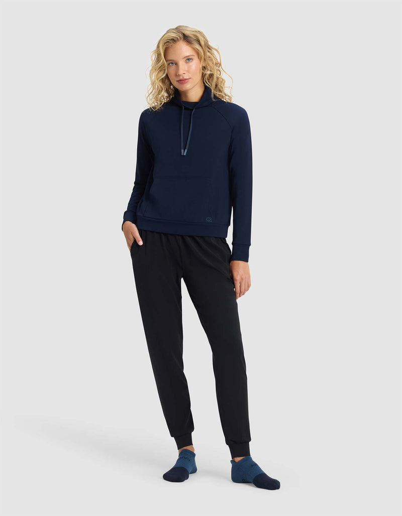 A woman with curly blonde hair wears the Cozy Earth Women's Brushed Bamboo Mock Neck, paired with black jogger pants and navy socks, standing against a plain light gray background with a relaxed posture and a slight smile.