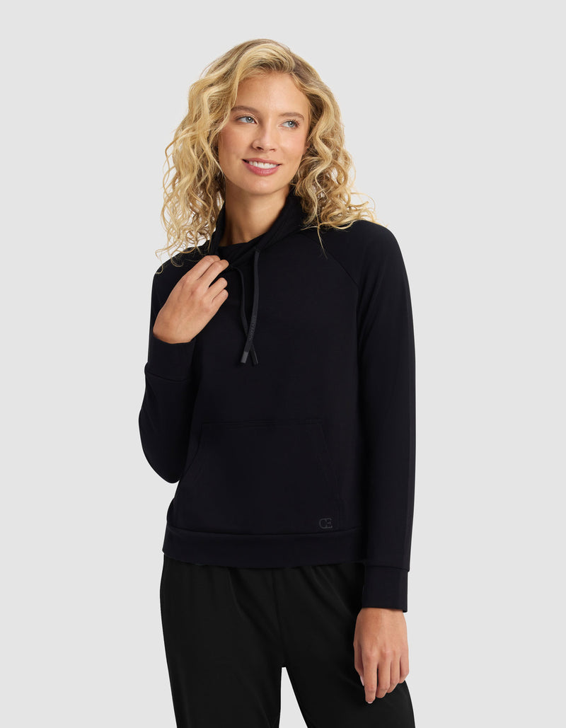 A woman with curly blonde hair wears the Cozy Earth Women's Brushed Bamboo Mock Neck & Jogger Set in black. She smiles slightly, looks to the side, and touches the drawstring. The background is plain light gray.
