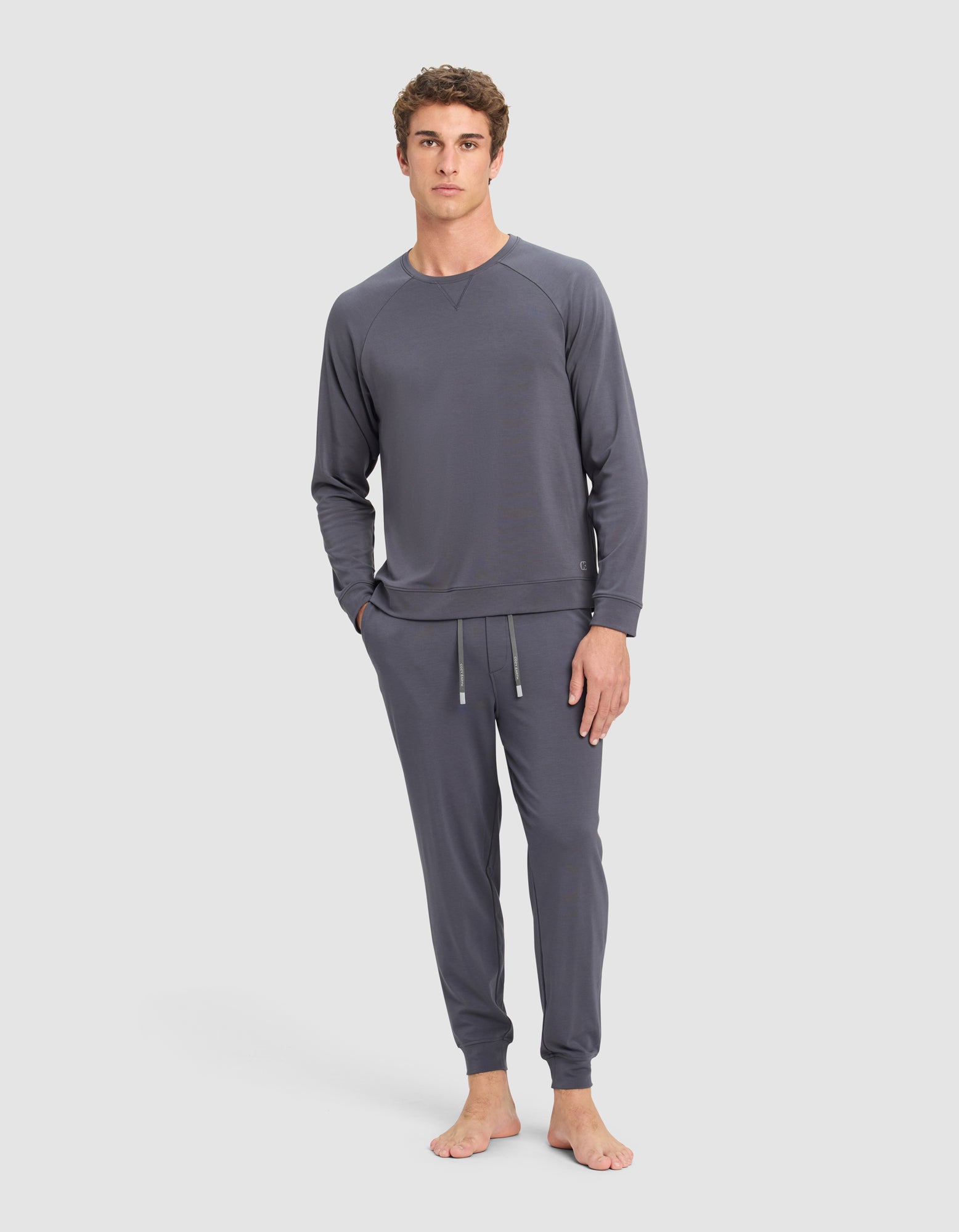 A man with short curly hair and a relaxed expression stands barefoot against a plain background, wearing the Cozy Earth Men's Brushed Bamboo Jogger Set in slate gray, featuring a long-sleeve top and drawstring jogger-style pants.