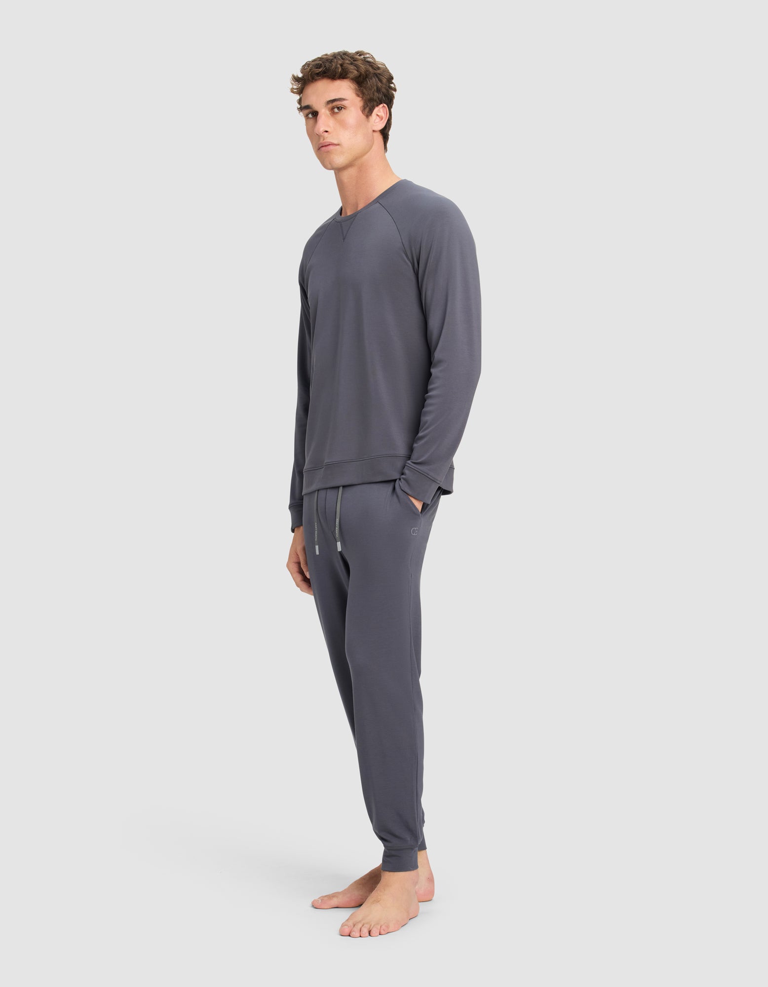 A man stands barefoot against a plain background, wearing the Cozy Earth Men's Brushed Bamboo Jogger Set in dark gray, featuring a matching long-sleeve top and jogger-style pants with his hands in his pockets.