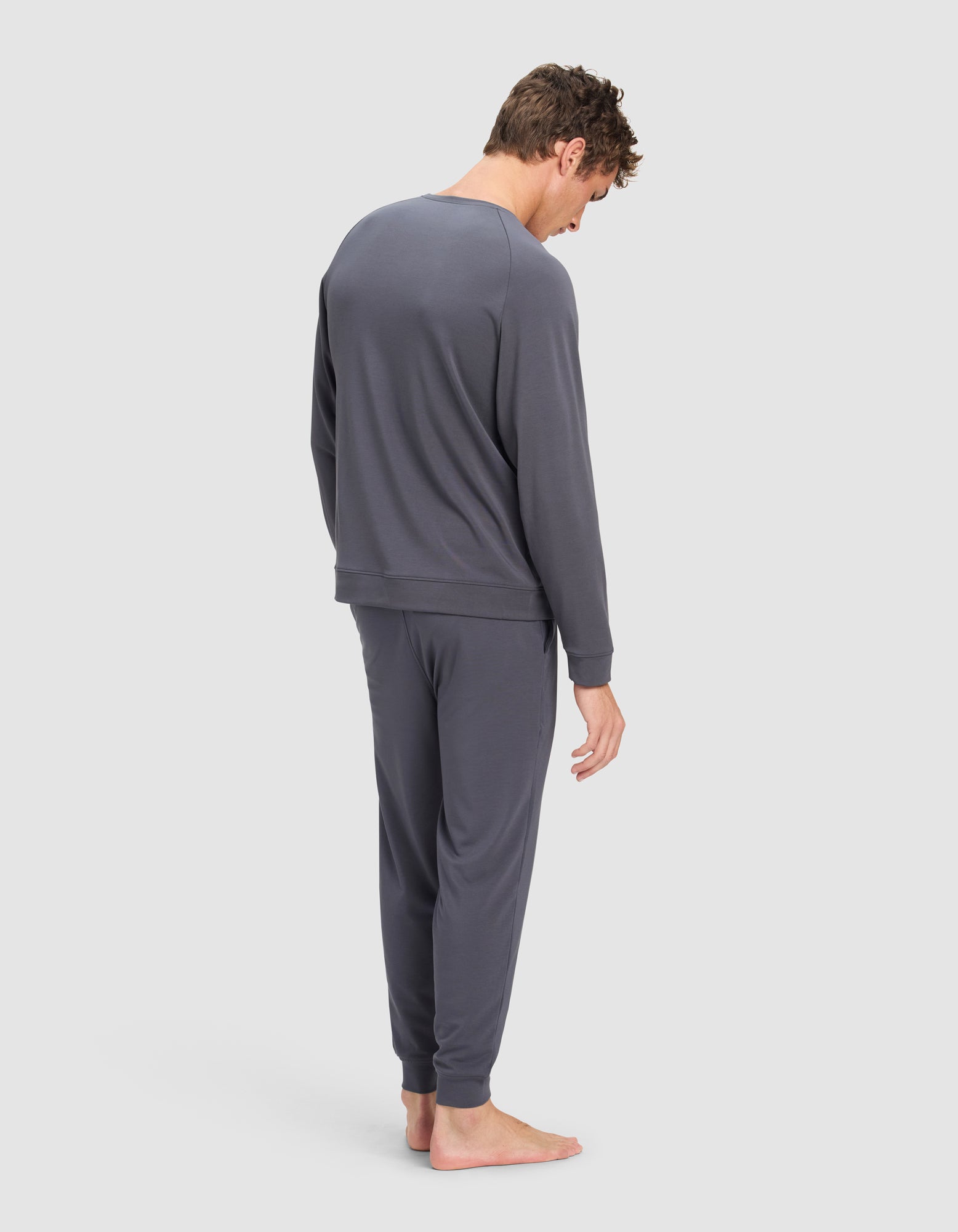 A man with short, tousled hair stands barefoot facing away, wearing the Cozy Earth Men's Brushed Bamboo Jogger Set in dark gray, against a plain light background.