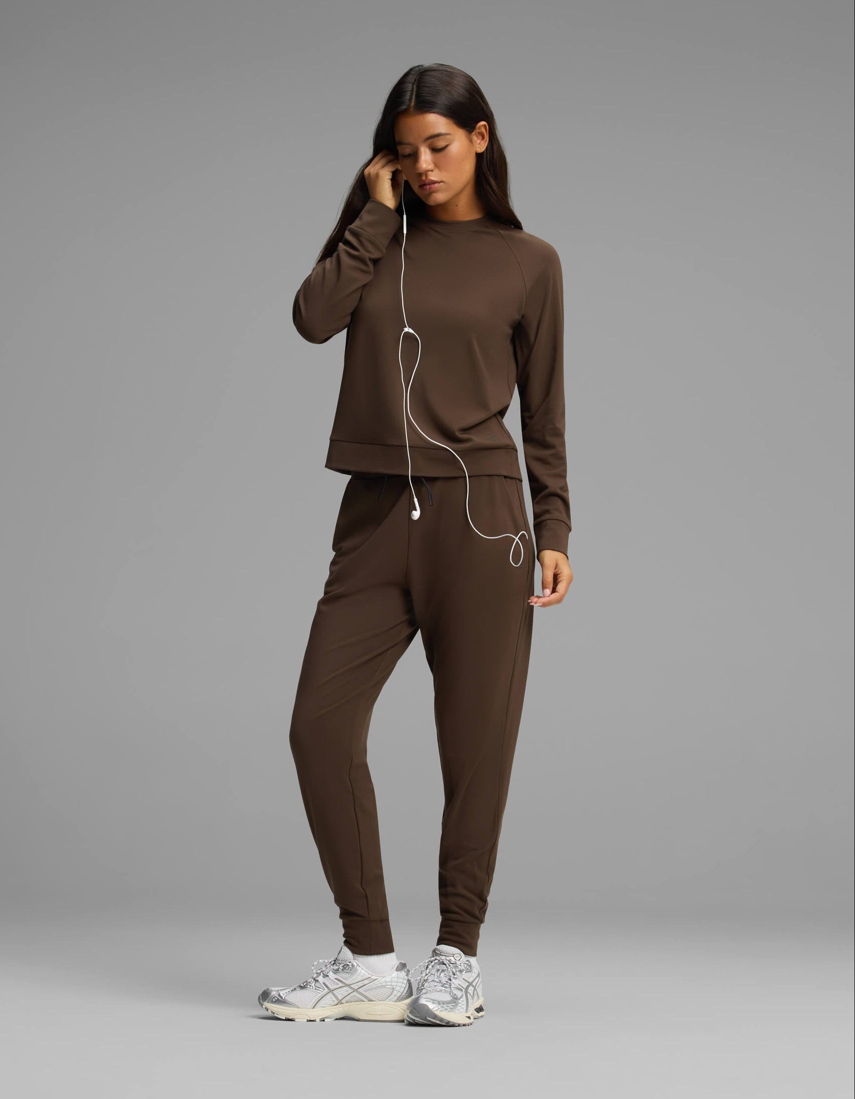 A woman wears the Cozy Earth Women's Brushed Bamboo Jogger Set, featuring a brown long-sleeve top and matching pants, as she stands in sneakers and holds white earphones to her ear against a plain gray background.