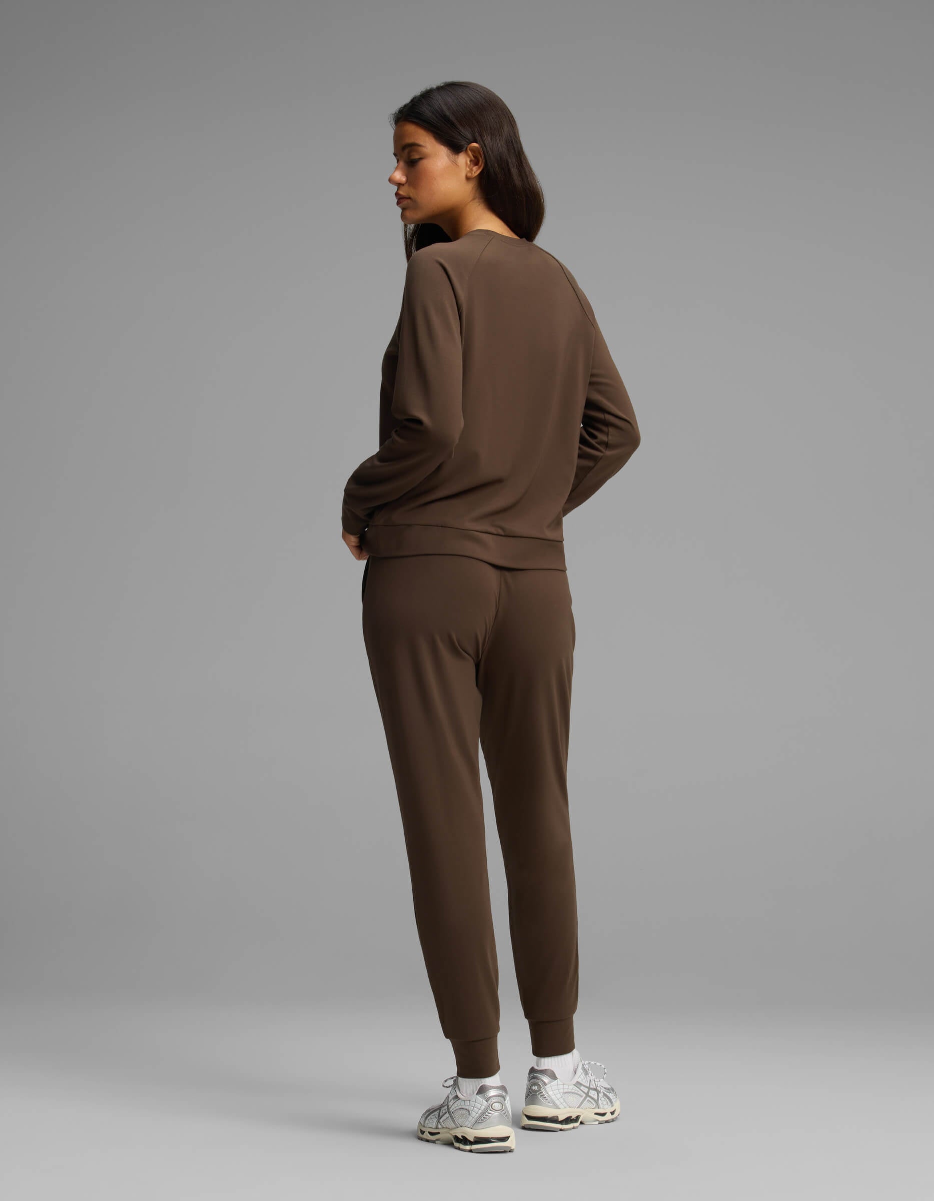 A woman models the Cozy Earth Women's Brushed Bamboo Jogger Set, featuring a brown long-sleeve top and matching jogger pants. She faces away and glances to her left, wearing white sneakers against a plain gray background.