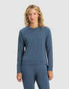 A woman with curly blonde hair faces forward against a plain light gray background, wearing the Cozy Earth Women's Brushed Bamboo Pullover Crew in blue, paired with matching pants.|Color:Dawn