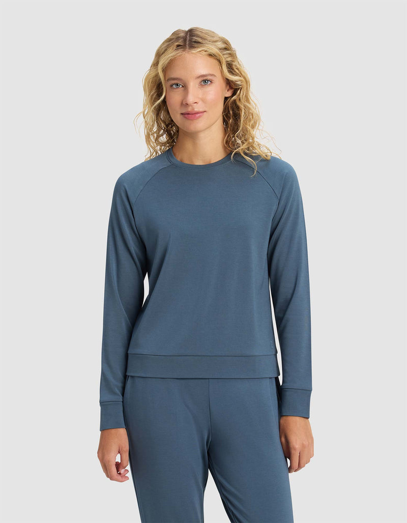 A woman with curly blonde hair faces forward against a plain light gray background, wearing the Cozy Earth Women's Brushed Bamboo Pullover Crew in blue, paired with matching pants.