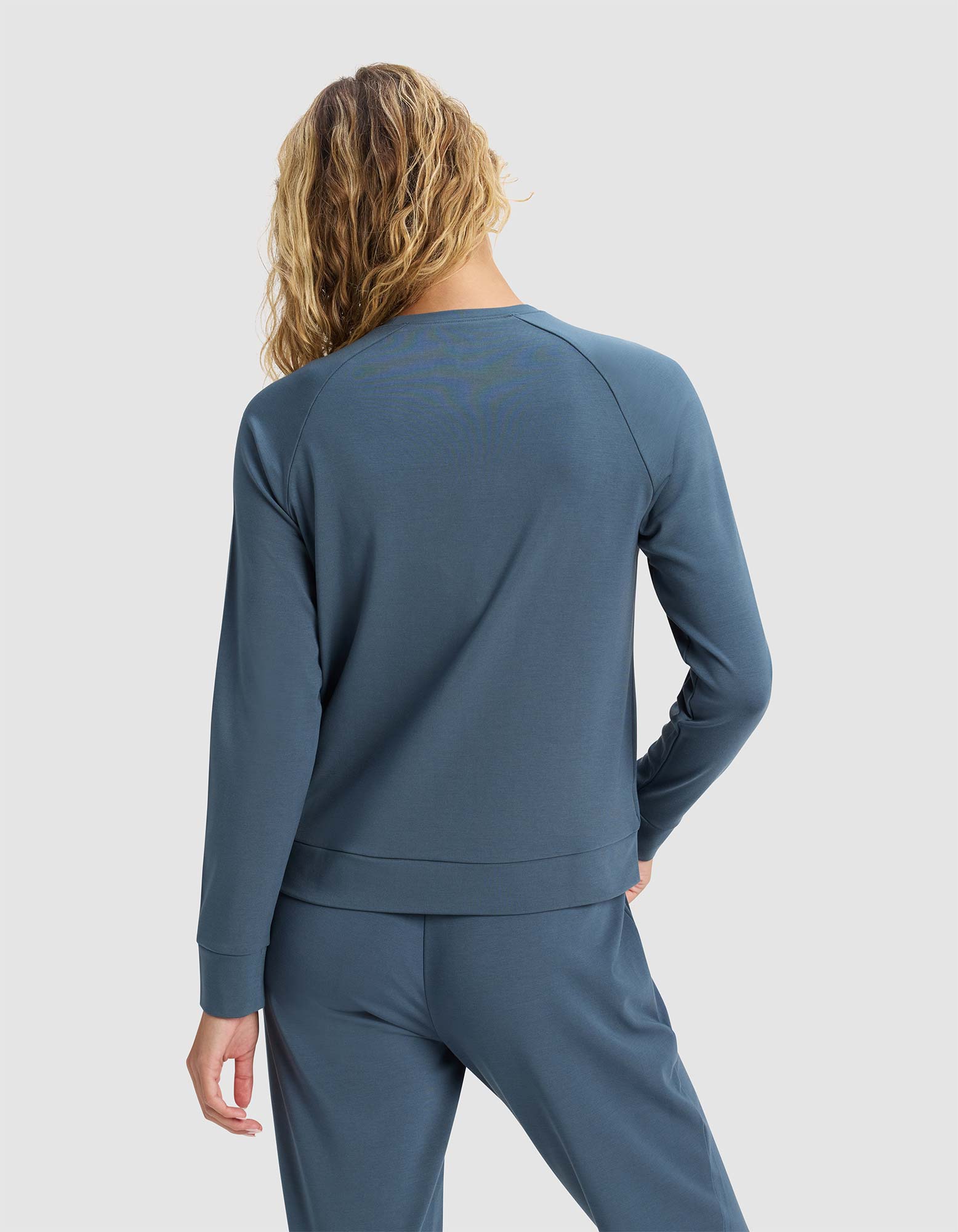 A woman with wavy, shoulder-length hair is seen from the back wearing the Cozy Earth Women's Brushed Bamboo Pullover Crew in blue. She stands against a plain, light background.