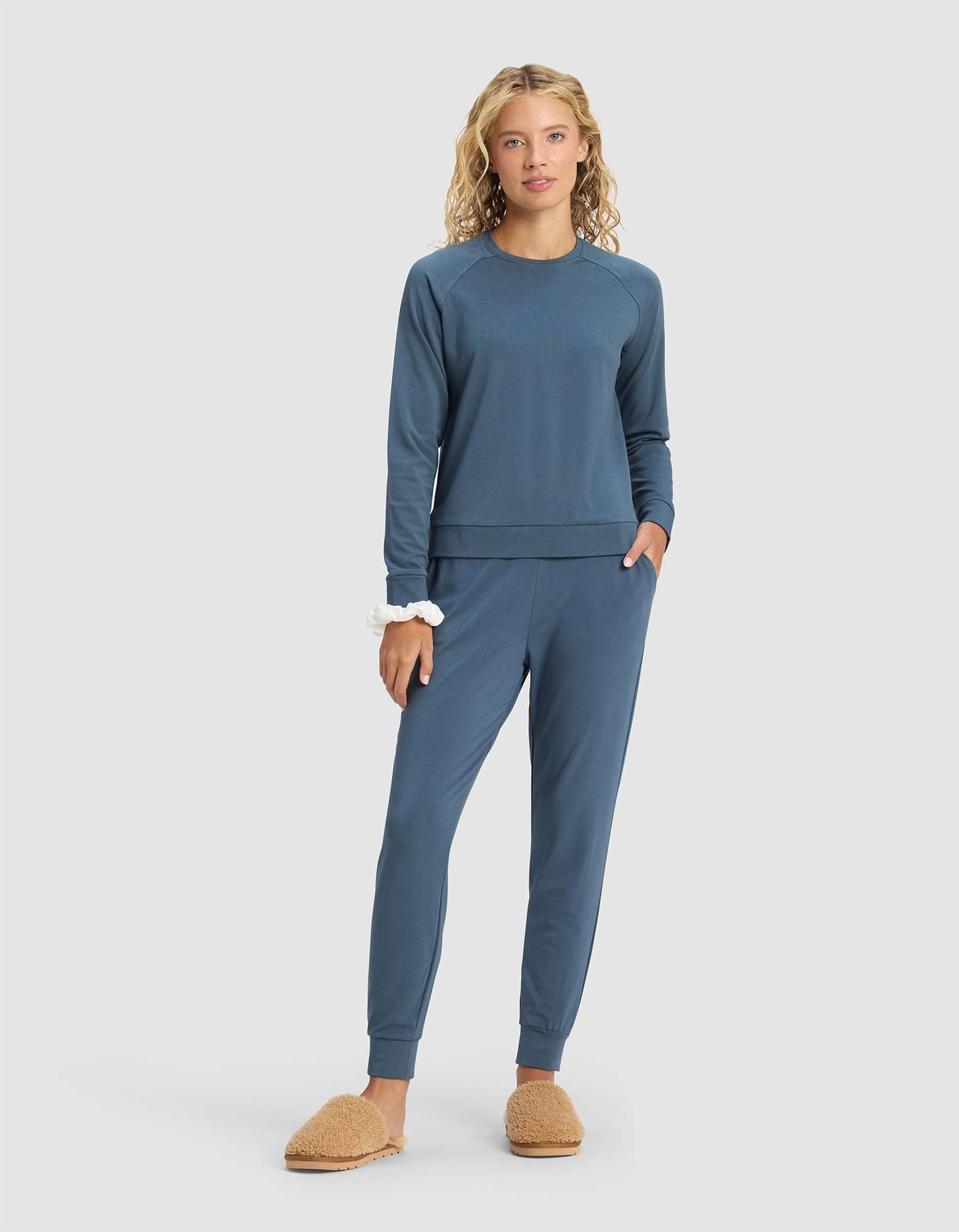 A woman faces forward against a plain light background, wearing the Cozy Earth Women's Brushed Bamboo Pullover Crew in blue with matching jogger pants, tan slippers, and a white scrunchie on her wrist. 